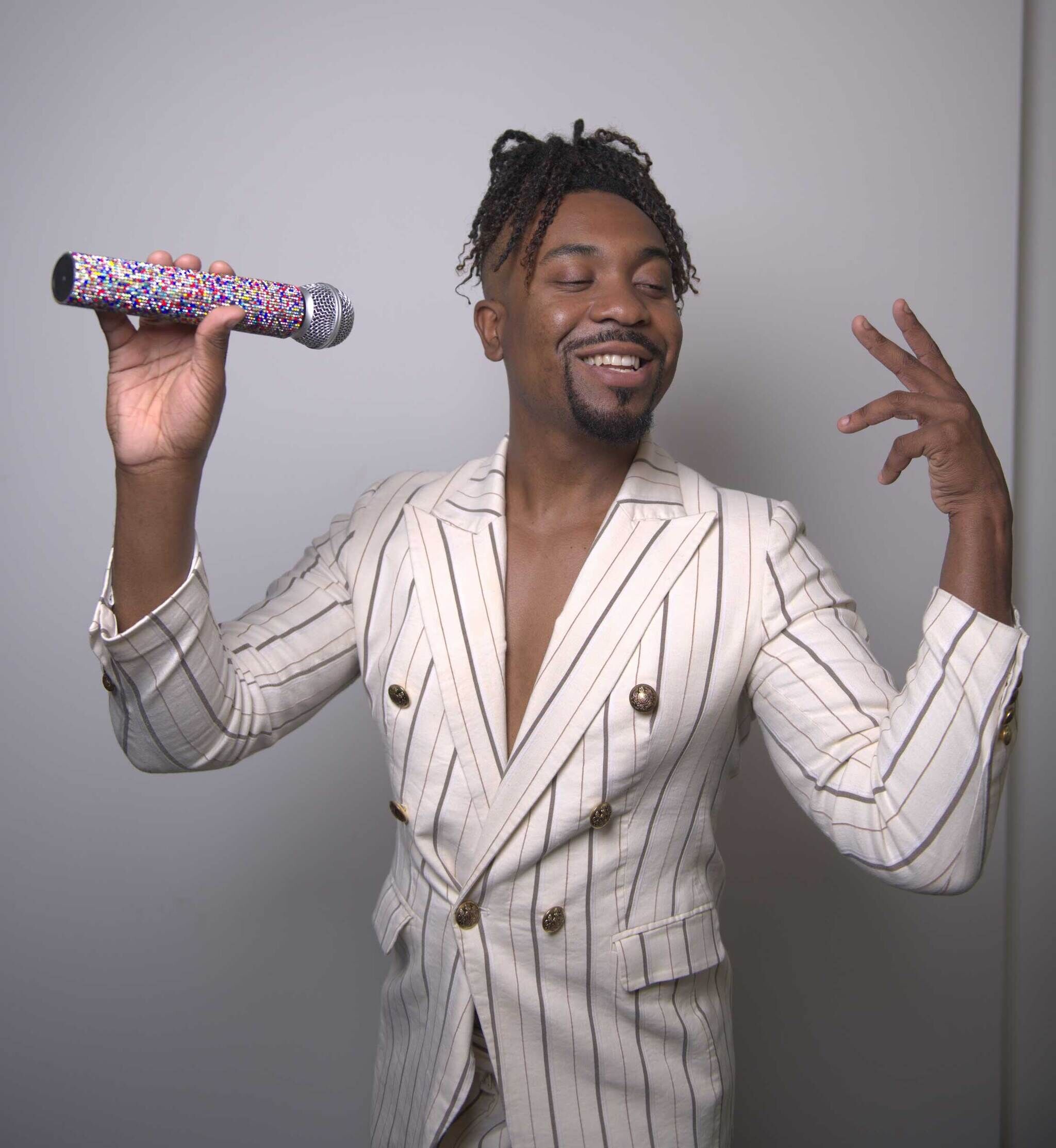 A smiling African American vocalist with his head slightly turned and eyes shut radiates joy. The vocalist is wearing a pin-striped blazer without a shirt underneath and has a braided updo slightly framing his face. He is posing with a multi-colored bedazzled microphone in one hand and the other is in an artistic hand gesture.