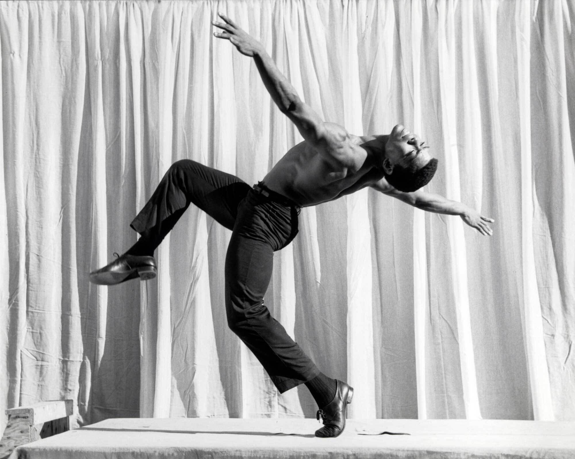 A shirtless man, Alvin Ailey, in black pants and shoes performs a dynamic dance pose against a backdrop of white curtains.