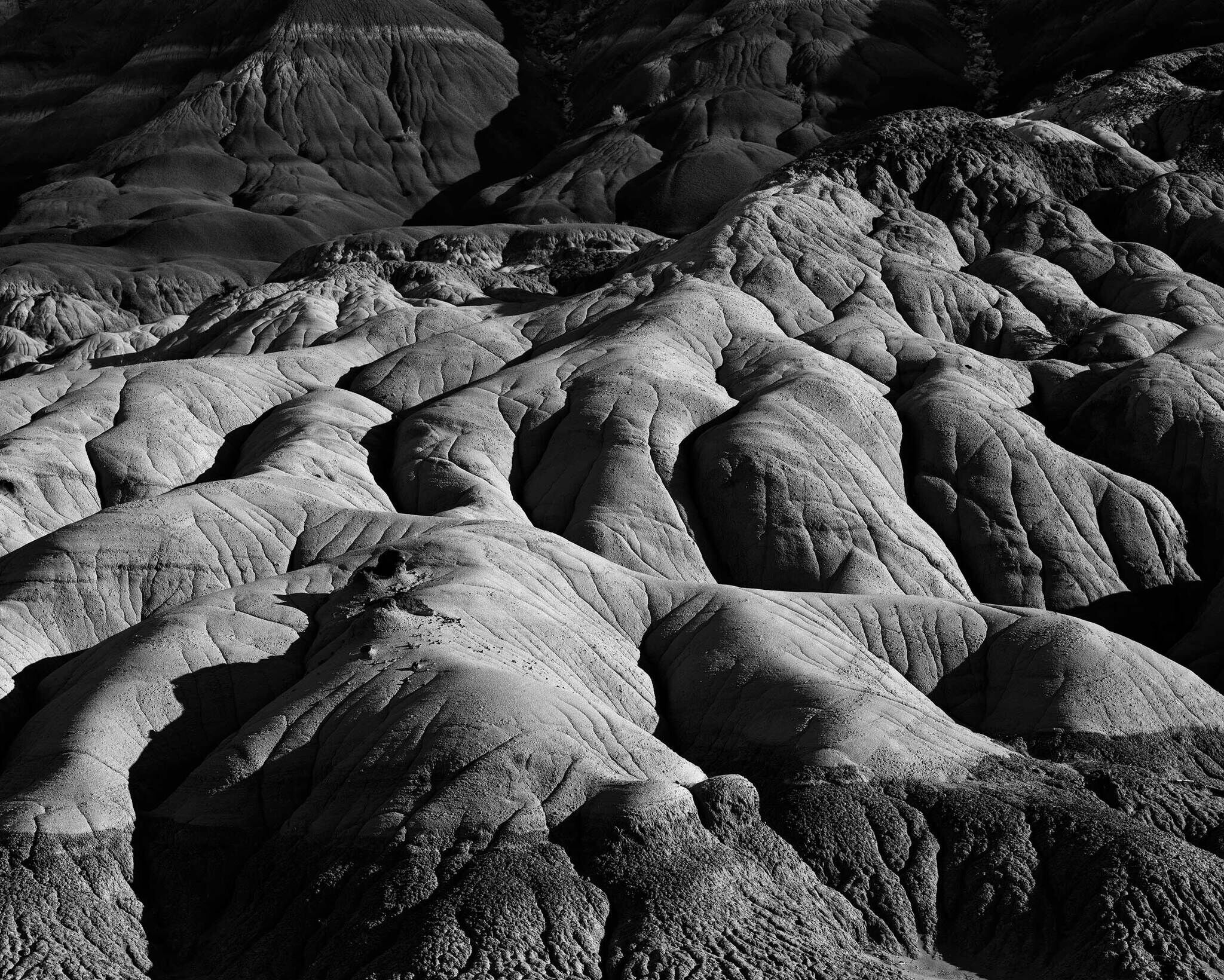 A black and white photograph of eroded, textured rock formations resembling waves or folds, creating a dramatic and rugged landscape.