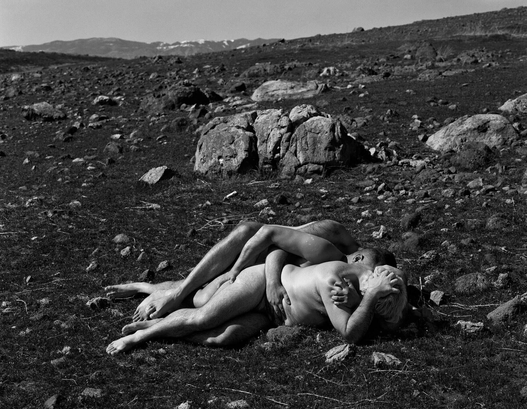 A black and white photograph of two people lying intertwined on a rocky, barren landscape with mountains in the background.