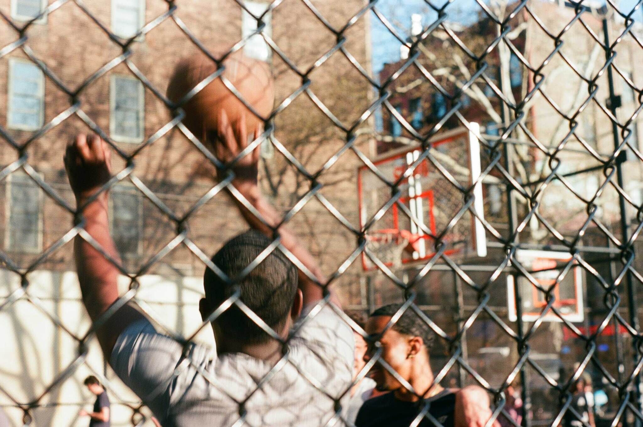 Two people playing basketball on an outdoor court, viewed through a chain-link fence. One person is shooting the ball towards the hoop.