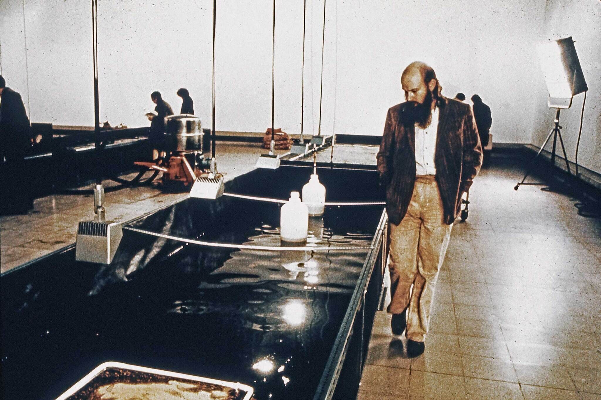 A man with a beard walks beside a series of water tanks in a gallery, with other people in the background.