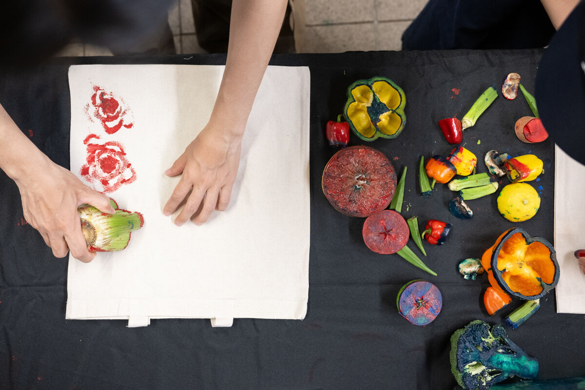Hands using cut vegetables dipped in paint to stamp patterns on fabric; various painted vegetables scattered on a black tablecloth.