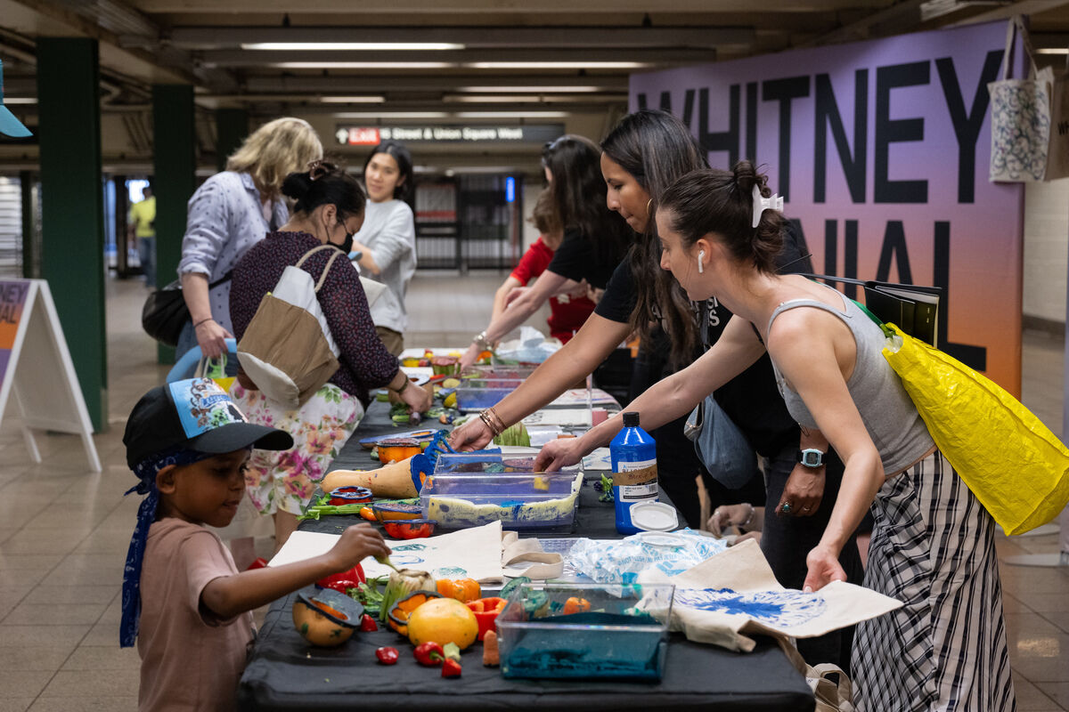 People engaging in an art activity at a table in a subway station, with a large "Whitney" sign in the background.