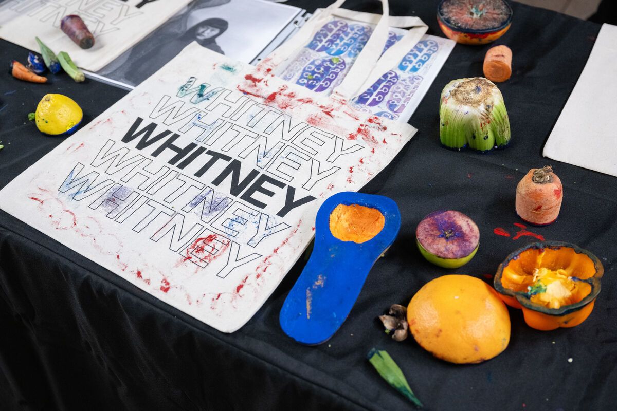 Canvas bag with "WHITNEY" printed multiple times, surrounded by colorful fruits and vegetables on a black tablecloth.