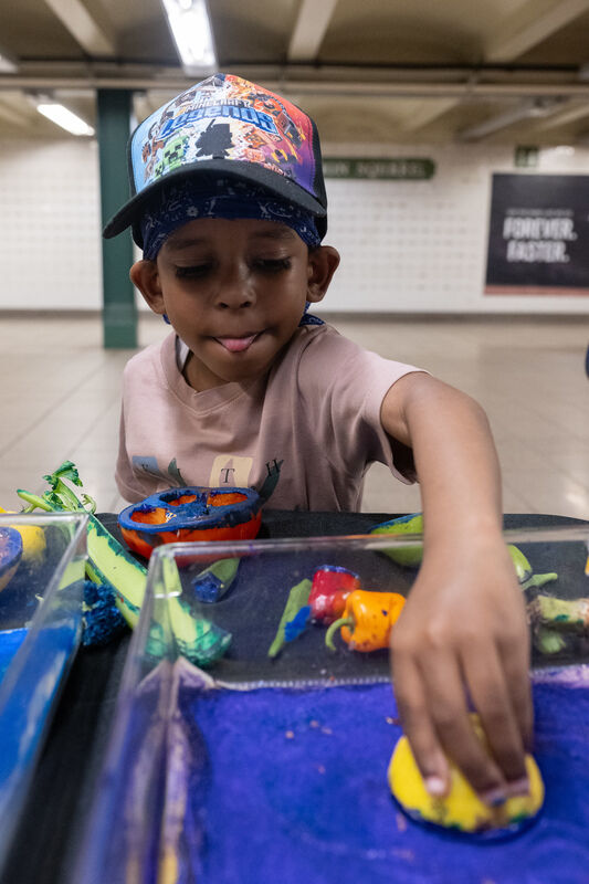 A child wearing a colorful cap and bandana plays with paint, dipping a sponge into blue paint on a table in a spacious indoor area.
