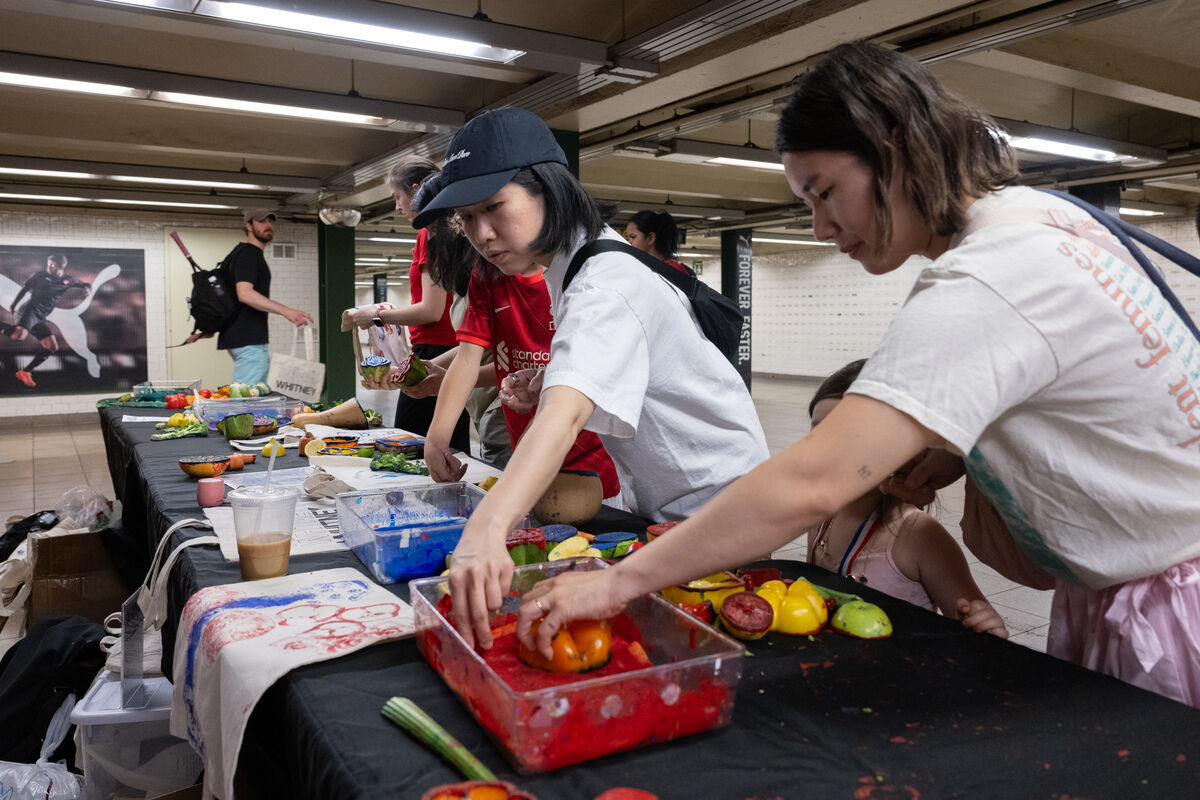 People at a table in a subway station, engaging in an art activity with colorful paints and vegetables. A large sports poster is visible in the background.