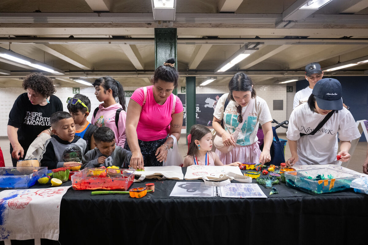 A group of children and adults engage in an art activity at a table in a subway station, using colorful paints and various tools.