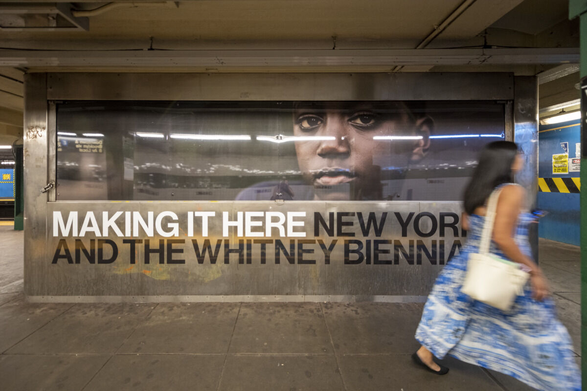 Subway station with a large poster showing a face and the text "MAKING IT HERE NEW YORK AND THE WHITNEY BIENNIAL." A woman in a blue dress walks by.
