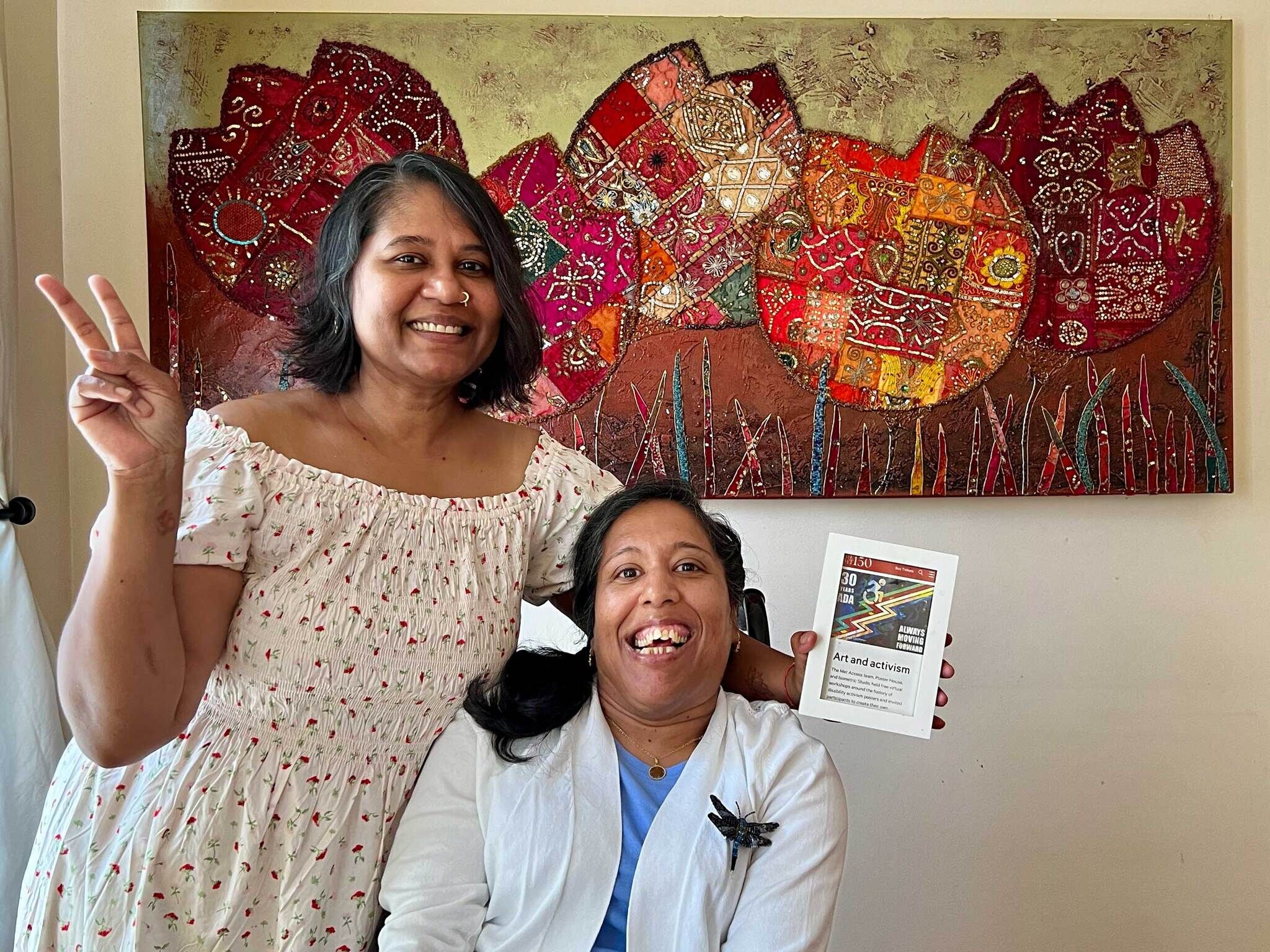 Lakshmee and Annie are Indo-Caribbean, women of color and mid age. Lakshmee has non apparent disabilities and Annie, who uses a wheelchair, has visible disabilities. They’re standing in front of a very colorful painting. Lakshmee hugs Annie from the back of the wheelchair and are both smiling for the camera. One of Lakshmee's hand showcase the peace sign while the other that embraces Annie holds up a photo of a Disability Pride flag.