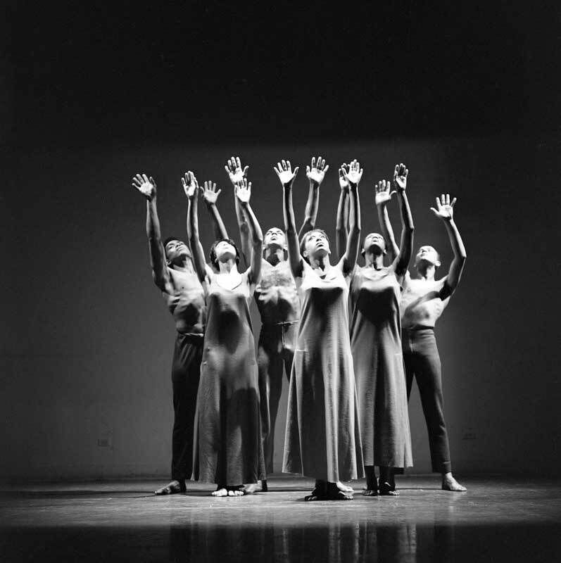 Black and white photograph of a dance performance with three male and three female figures arranged in a group on stage, hands outstretched above. 