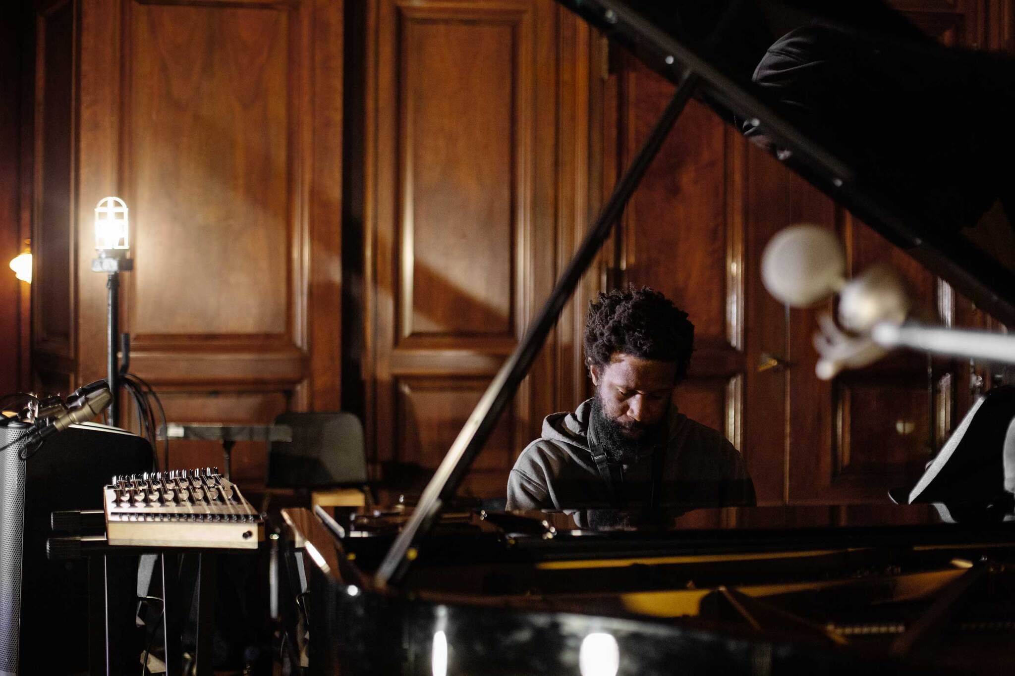 A portrait of JJJJJerome Ellis, a young Black person with short black hair and a black beard. Ellis sits at a grand piano in a room with dark wooden walls surrounded by musical instruments. 