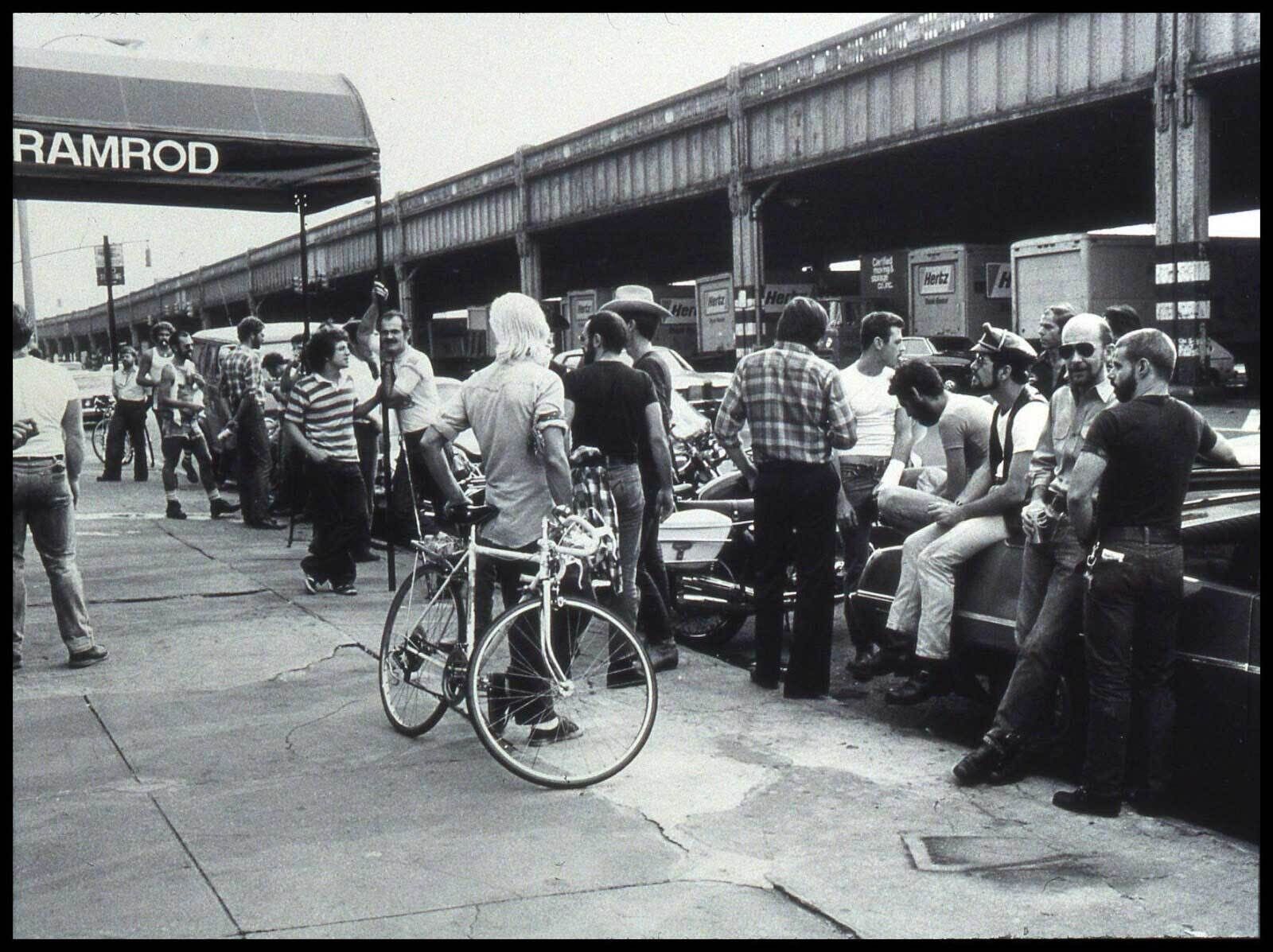Black and White photograph of a crowd of men casually leaning against parked cars or bicycles across from an elevated train rack. Part of an awning with block letters reading "Ramrod" is visible on the left.