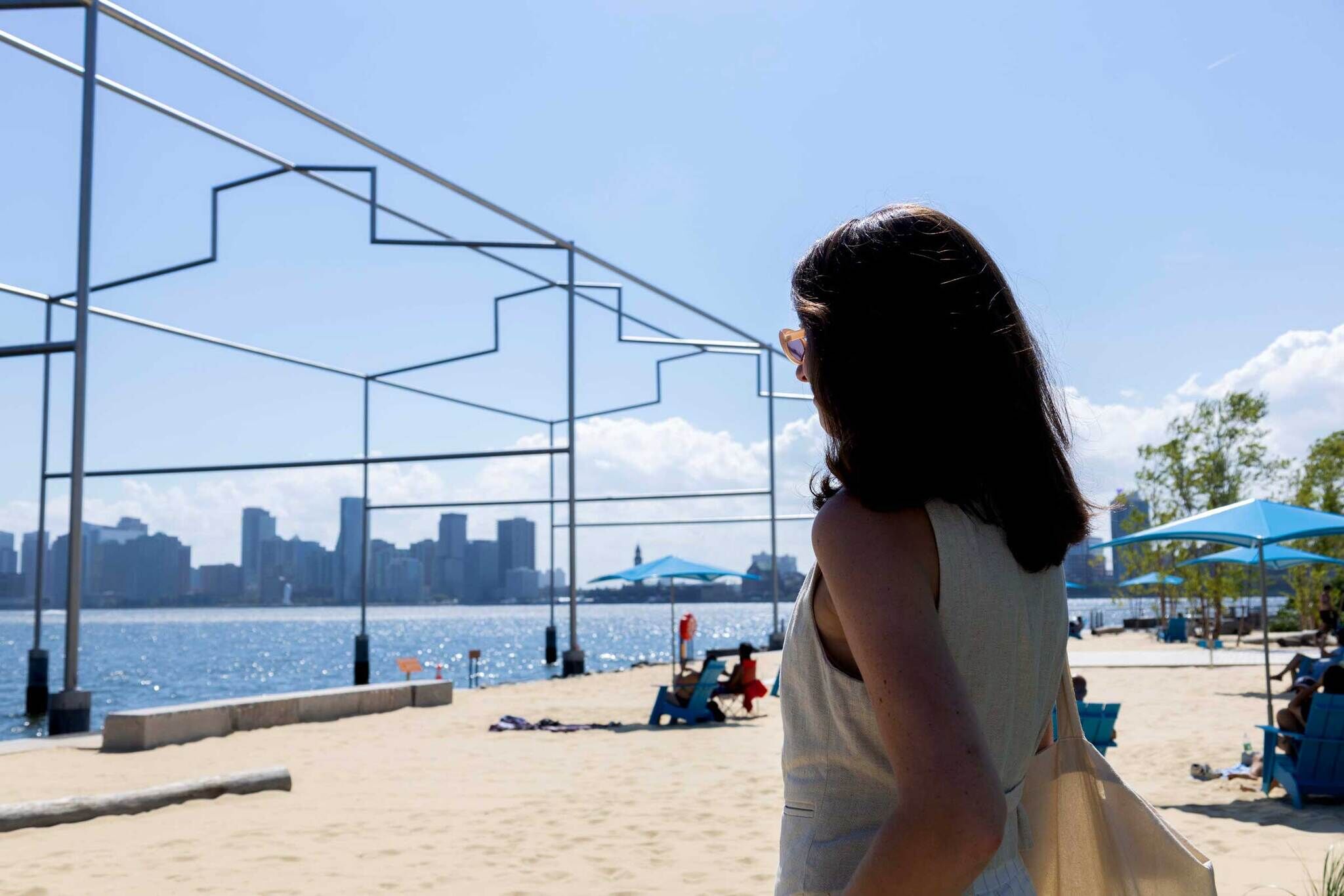 A woman with dark hair stands on a sandy beach, facing a city skyline across the water. Blue umbrellas and beach chairs are scattered around.