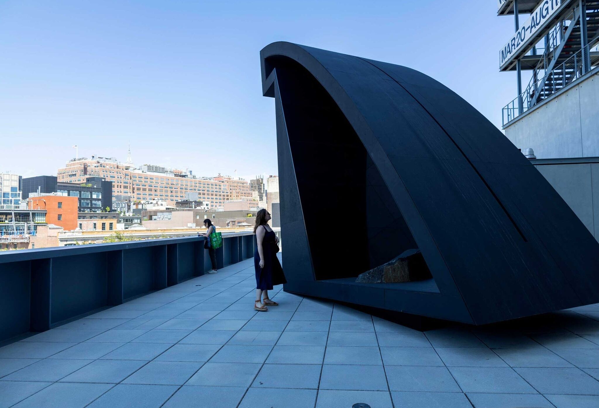 Two people stand on a rooftop terrace with a large, curved black sculpture. City buildings are visible in the background under a clear sky.