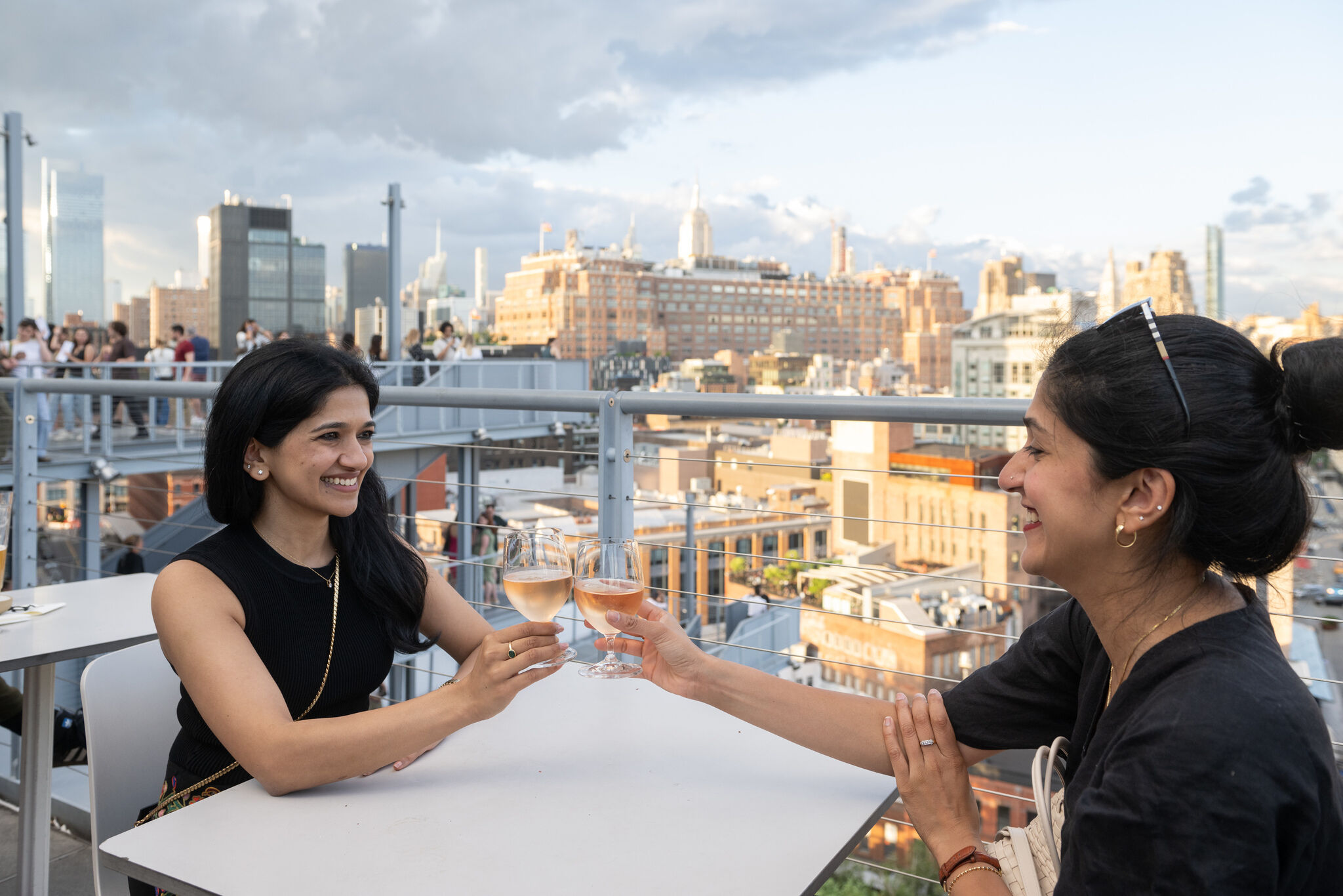 Two women clink glasses of rosé wine at a rooftop bar with a cityscape in the background.
