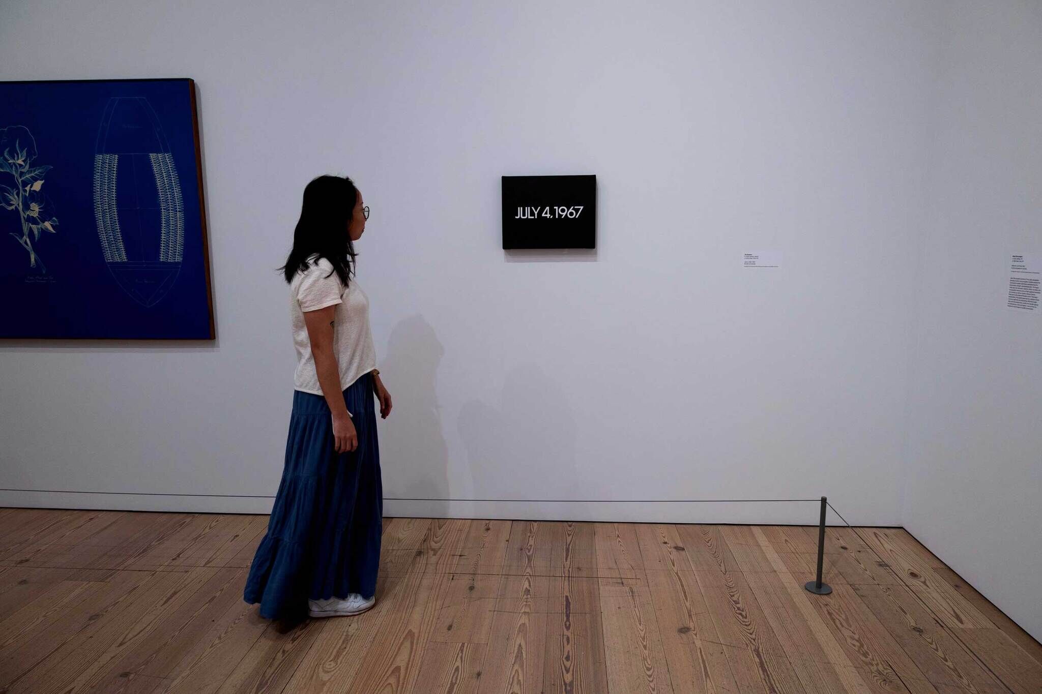 A woman in a white shirt and blue skirt looks at a painting with "JULY 4, 1967" in a gallery. A blue artwork is on the adjacent wall.
