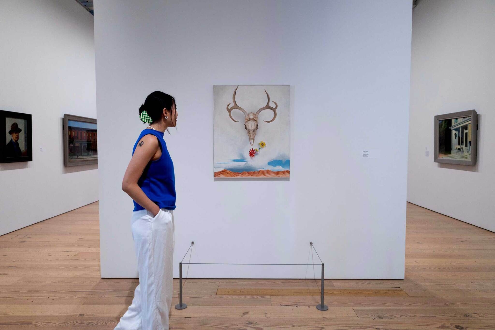 A woman in a blue top and white pants observes a painting of a skull with antlers in an art gallery. Other artworks are visible on the walls.