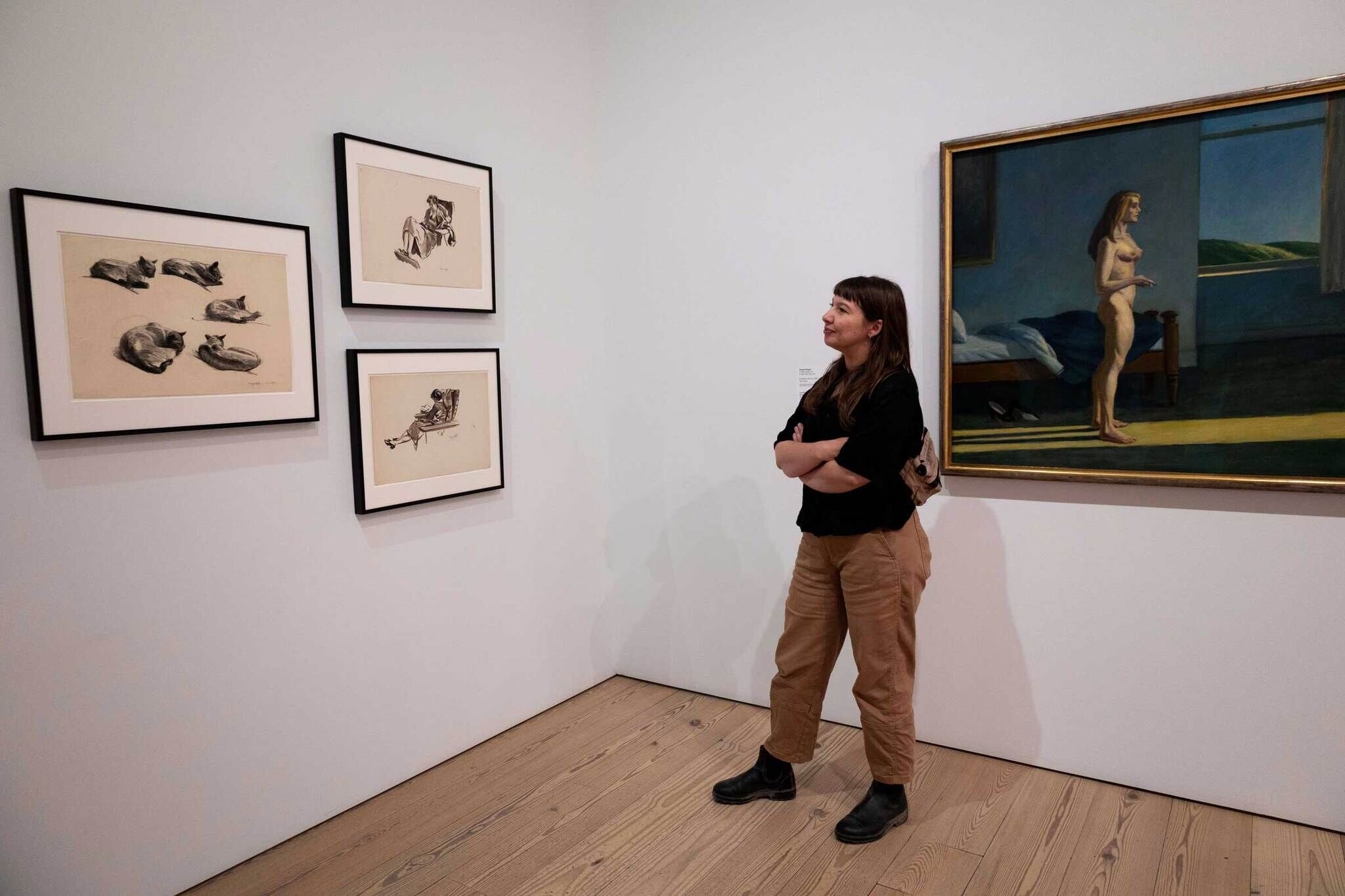 A woman stands in an art gallery, observing framed sketches of cats and people on the left wall and a painting of a nude woman by a window on the right.