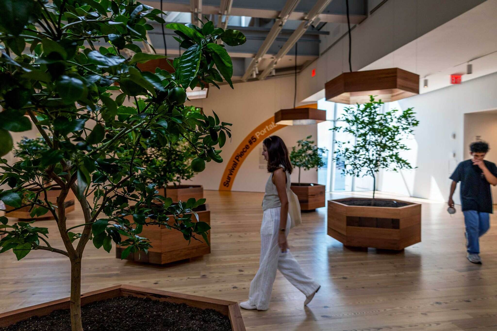 Two people walk through an indoor space with potted trees and a sign reading "Survival Piece #5 Portal."