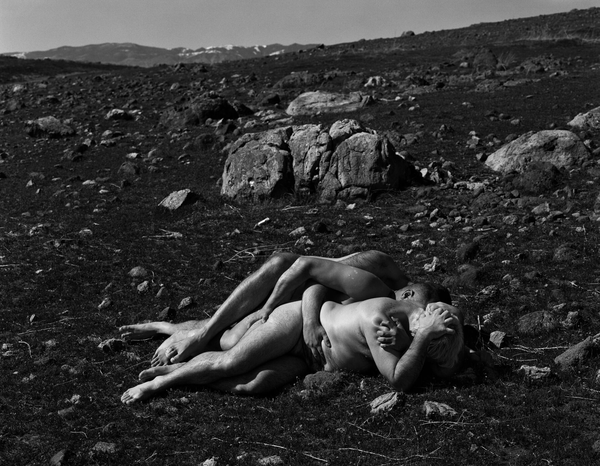 Two people embrace while lying on rocky terrain, with mountains in the background. The image is in black and white.
