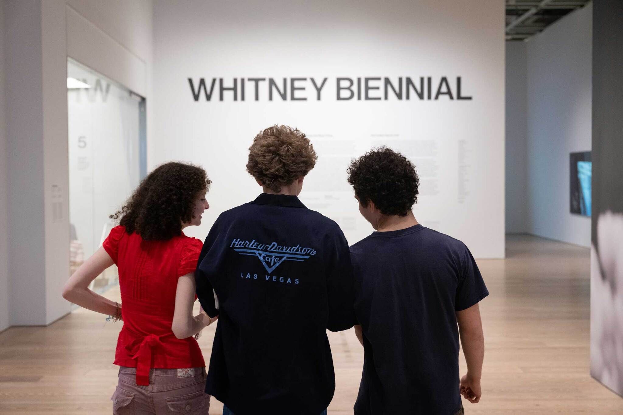 Three people stand in front of a wall with "Whitney Biennial" written on it, viewed from behind. One wears a red shirt, another a Harley-Davidson jacket.