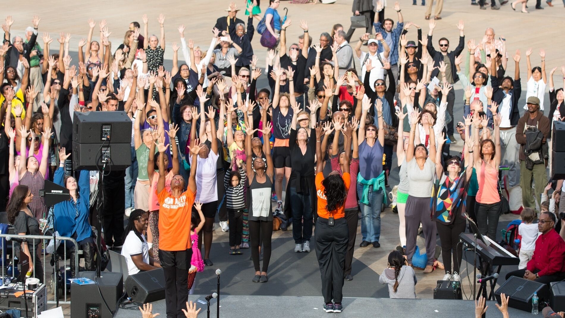 A large group of people stand with their arms raised in unison during an outdoor event, with a few individuals in orange shirts leading the activity.