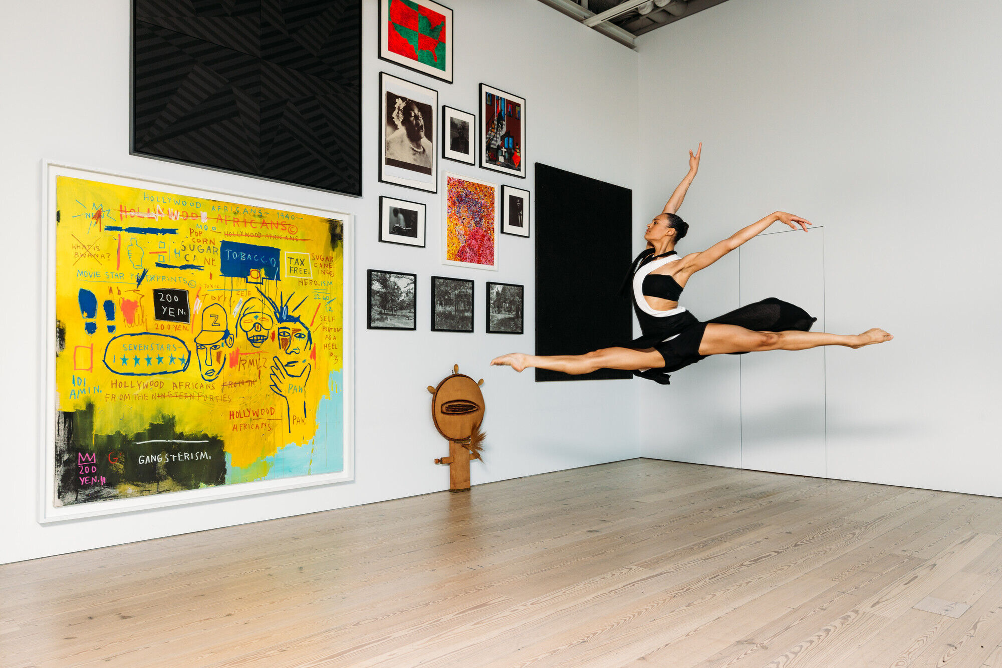 A dancer in a black and white outfit performs a grand jeté in an art gallery with colorful paintings and photographs on the walls.