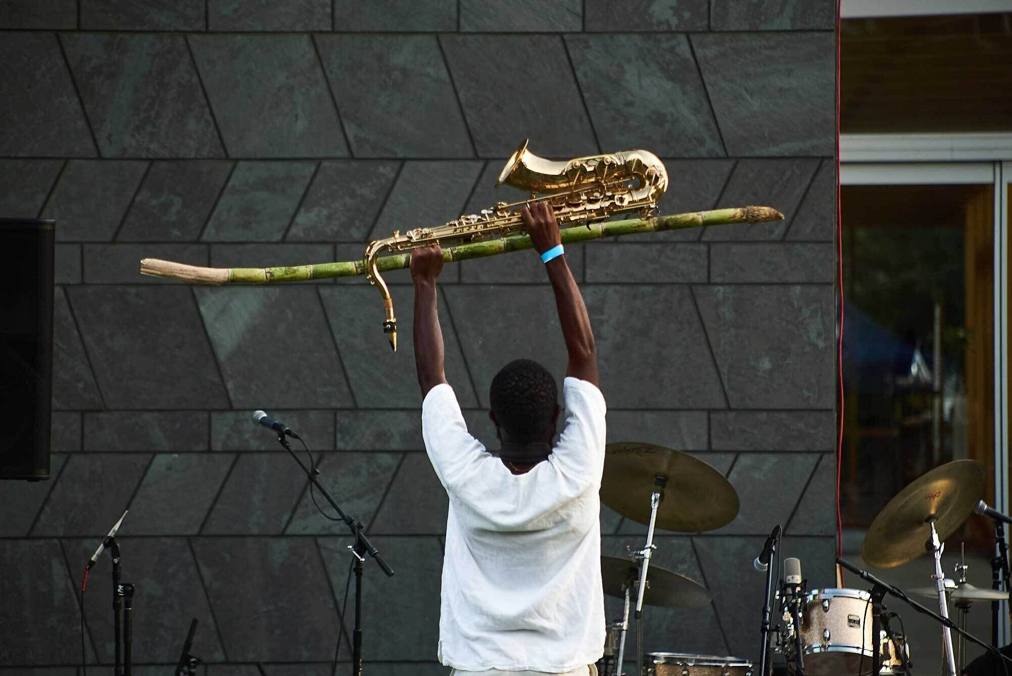 Person in a white shirt holds a saxophone and a long bamboo stick above their head on an outdoor stage with drums and microphones.