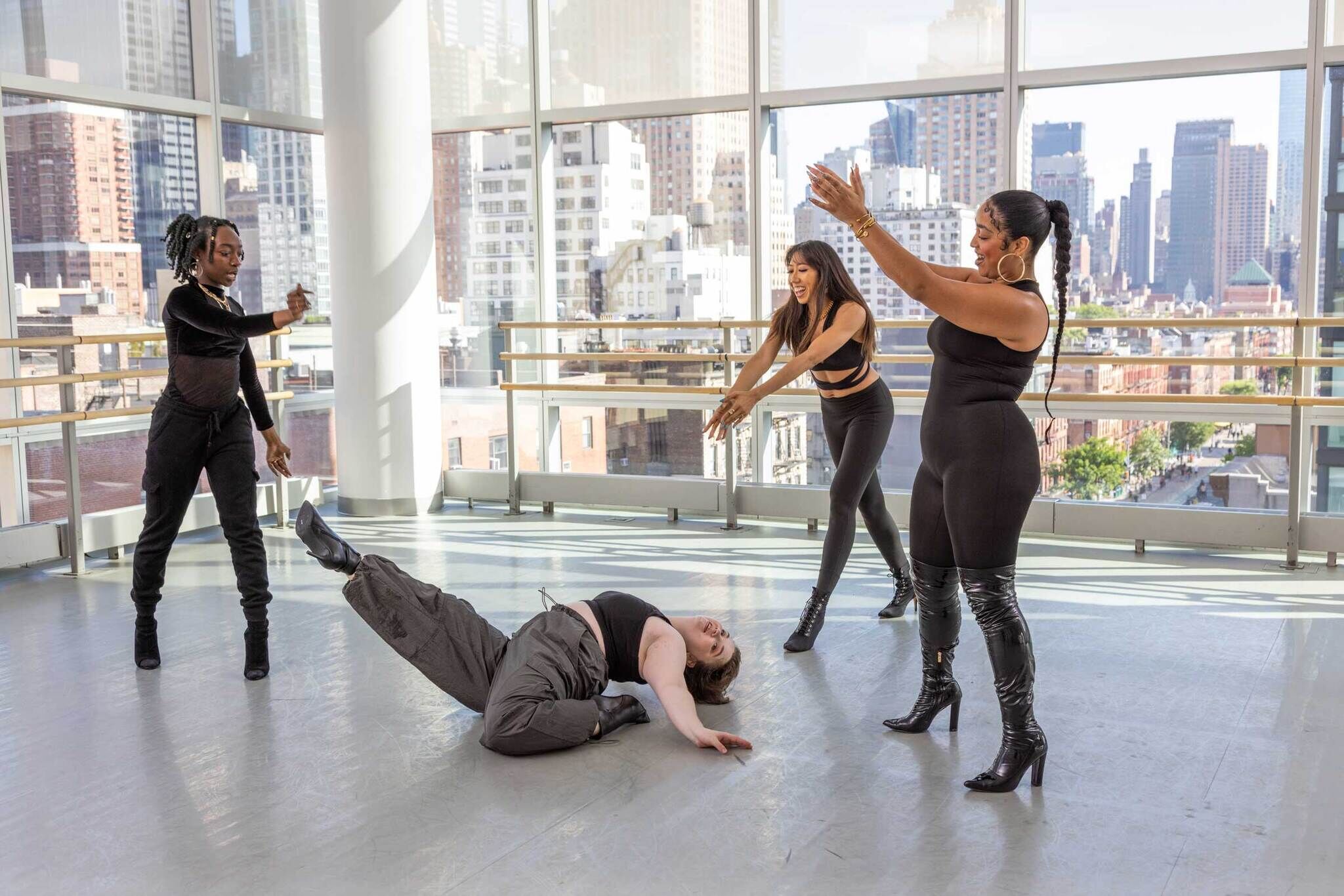 Four dancers practice in a studio with large windows and a city view. Three women stand and gesture toward a person doing a dip. She is has one leg bent, and the other in the air, and the head and torso are arched back onto the bent leg. All are wearing black dance attire and black heeled boots.