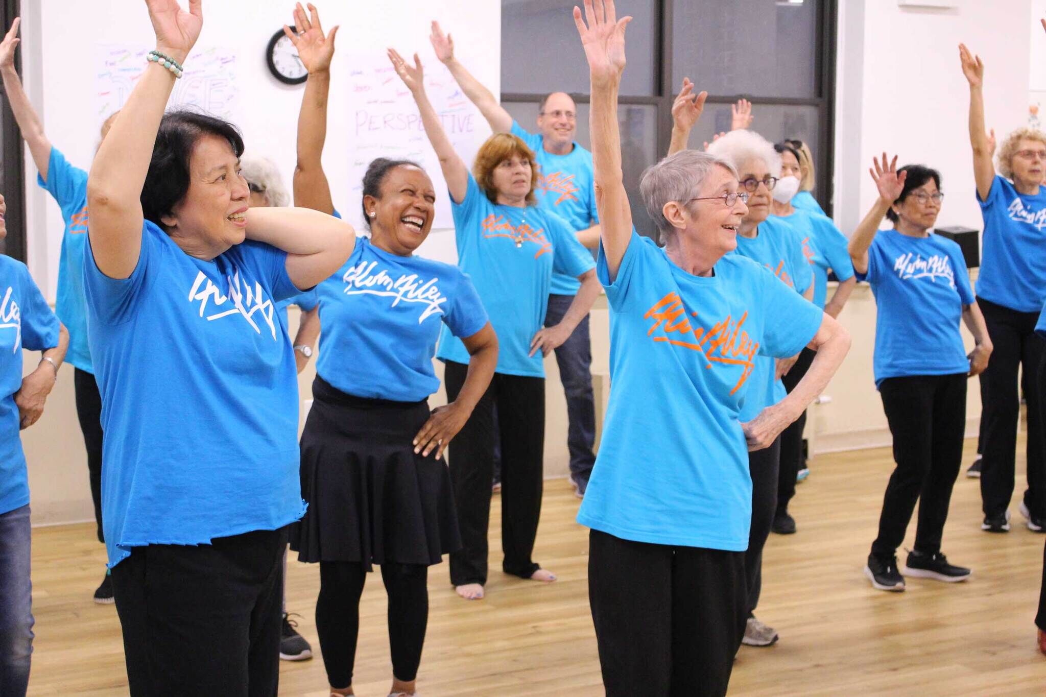 Group of older adults wearing blue ‘Alvin Ailey’ T-shirts learning a dance routine in an indoor dance studio.