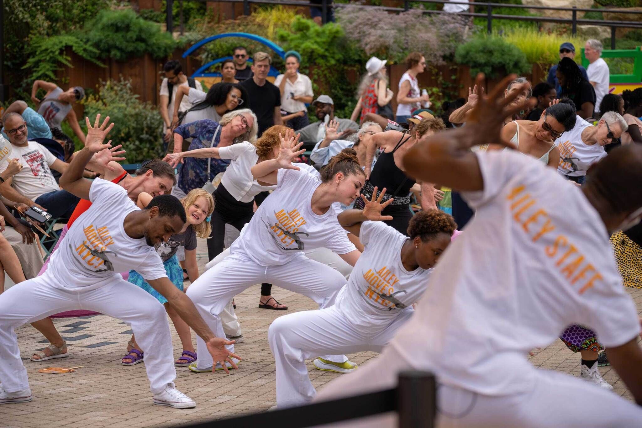 People wearing white “Ailey Staff” t-shirts teach a dance routine in an outdoor setting.