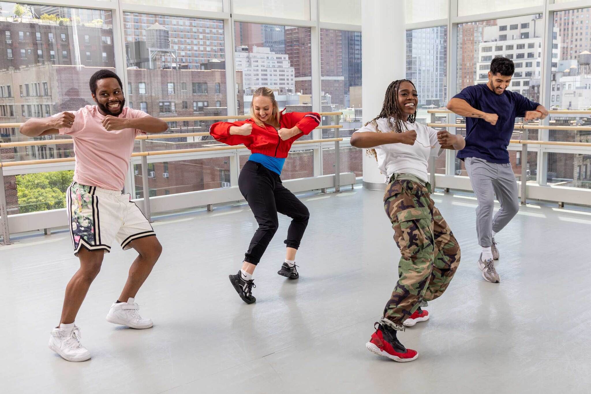 Four people dance energetically in a bright studio with large windows showing a cityscape. The group includes two men and two women dressed in athletic clothing. They smile and gesture dynamically, embodying a sense of fun, strength, and movement.