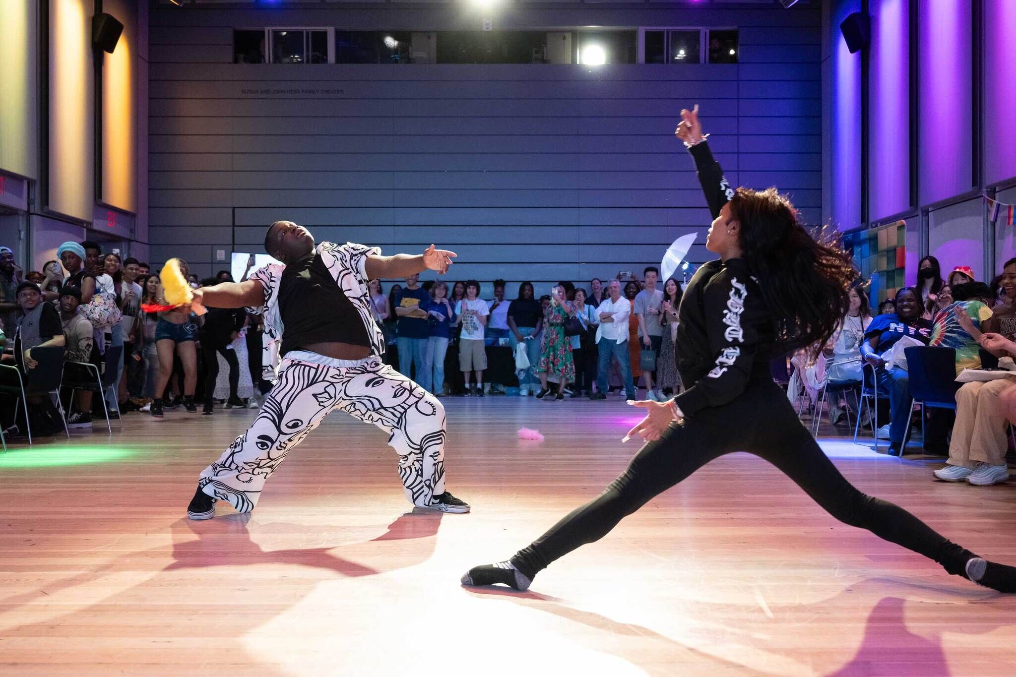 Two dancers perform energetically on a stage, with a crowd of people watching and cheering in the background.