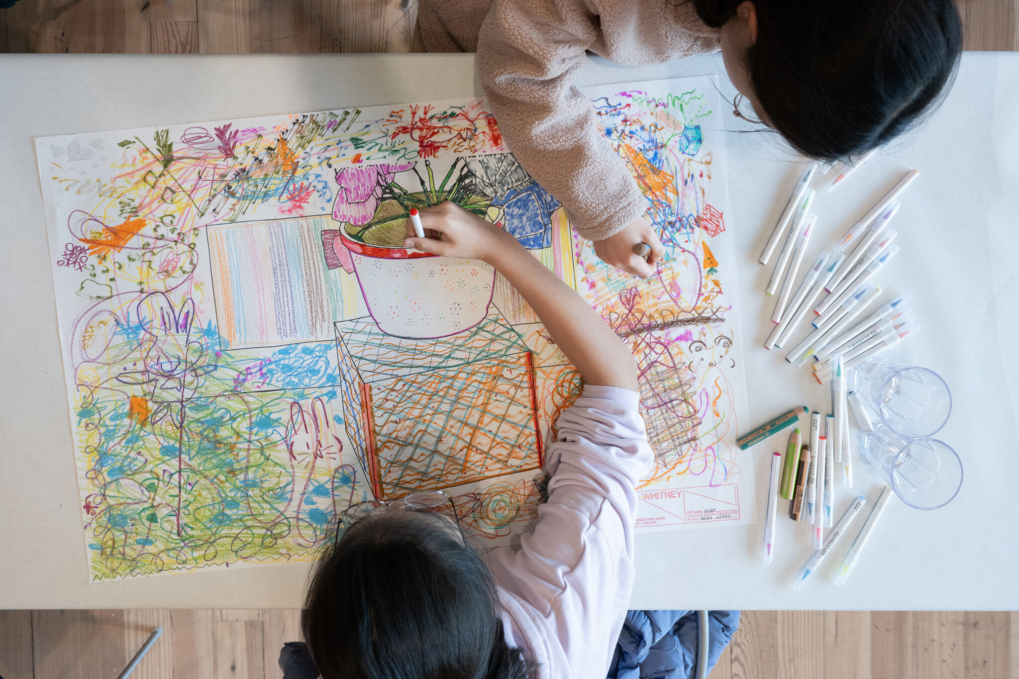 Two children drawing a colorful, abstract picture with markers on a large sheet of paper, surrounded by various art supplies.
