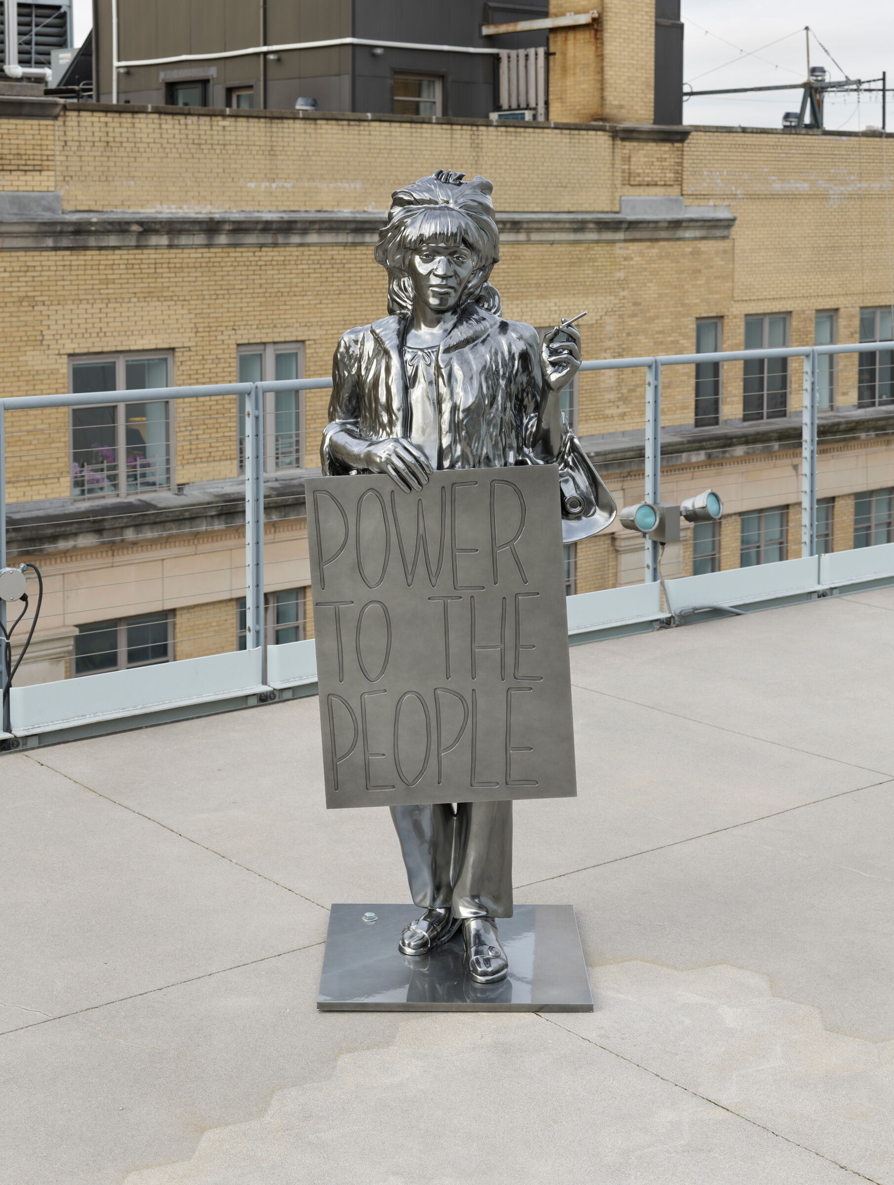 A metallic statue of a person holding a sign that reads "POWER TO THE PEOPLE" stands on a rooftop with a city building in the background.