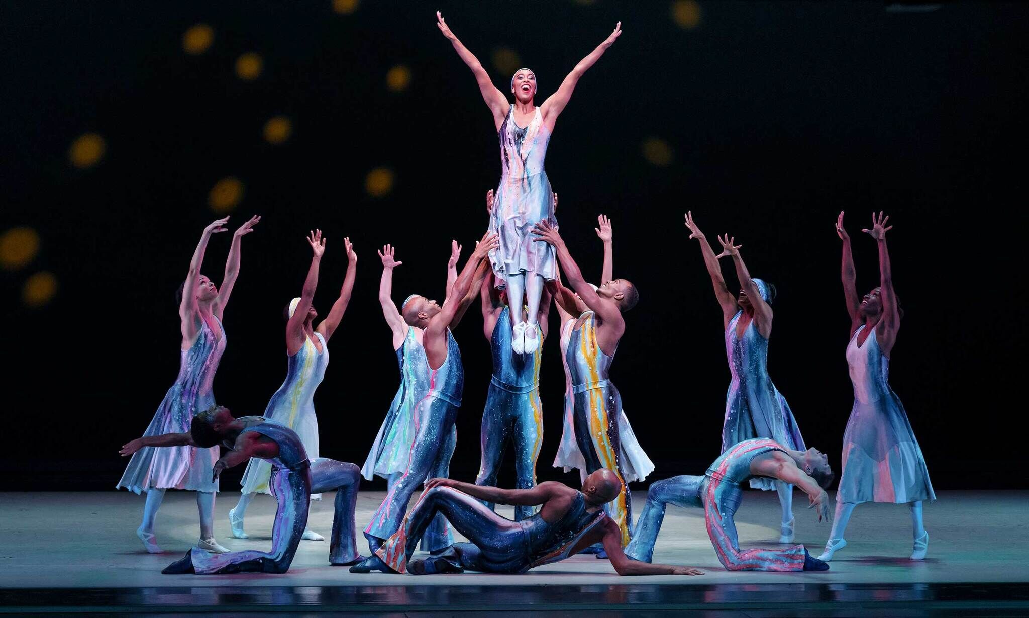 Ballet dancers in colorful costumes perform on stage, lifting one dancer high above the others with arms outstretched, against a dark background.