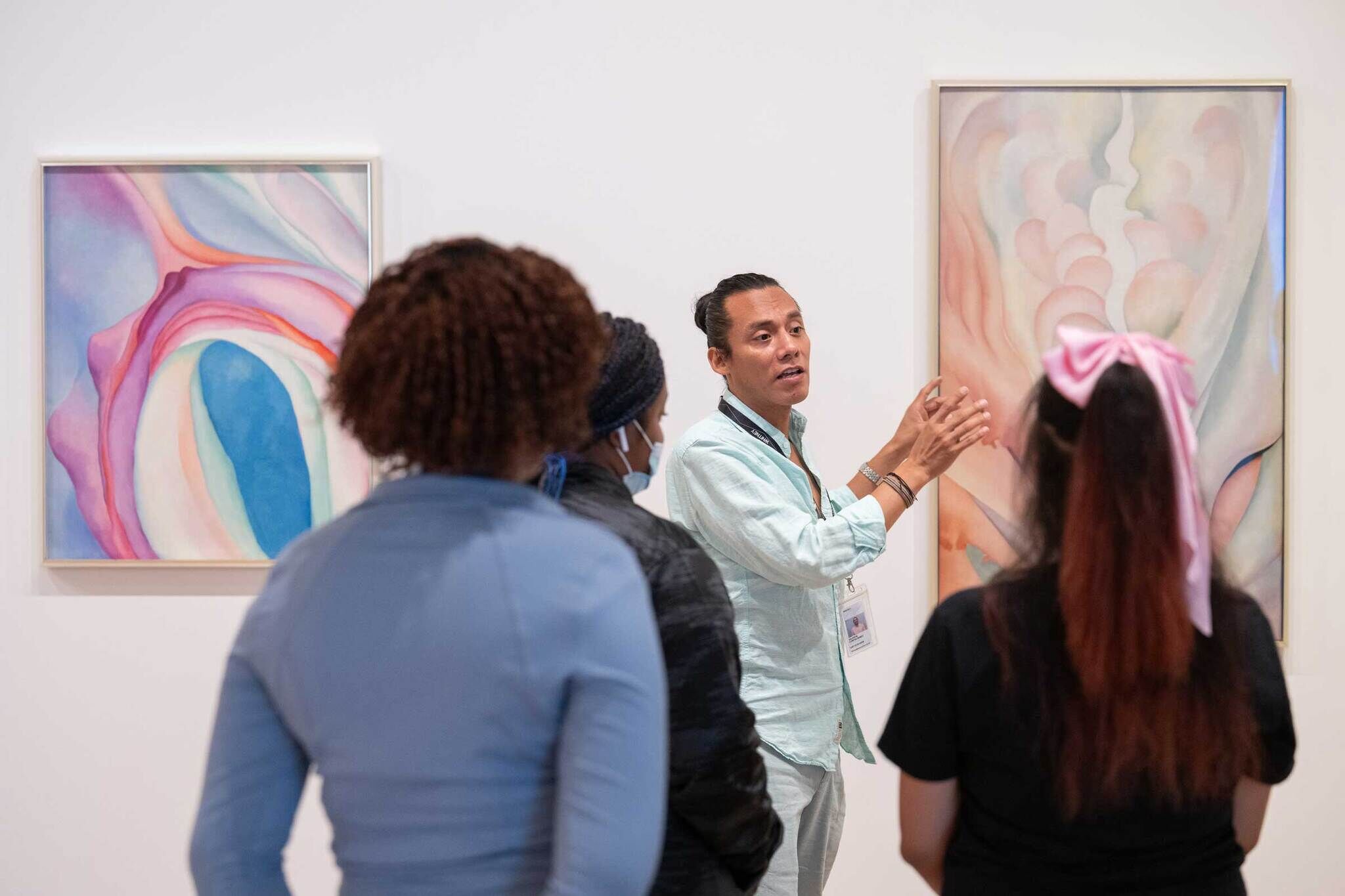 A guide explains abstract art to a group of three people in a gallery. Two colorful paintings are displayed on the wall.