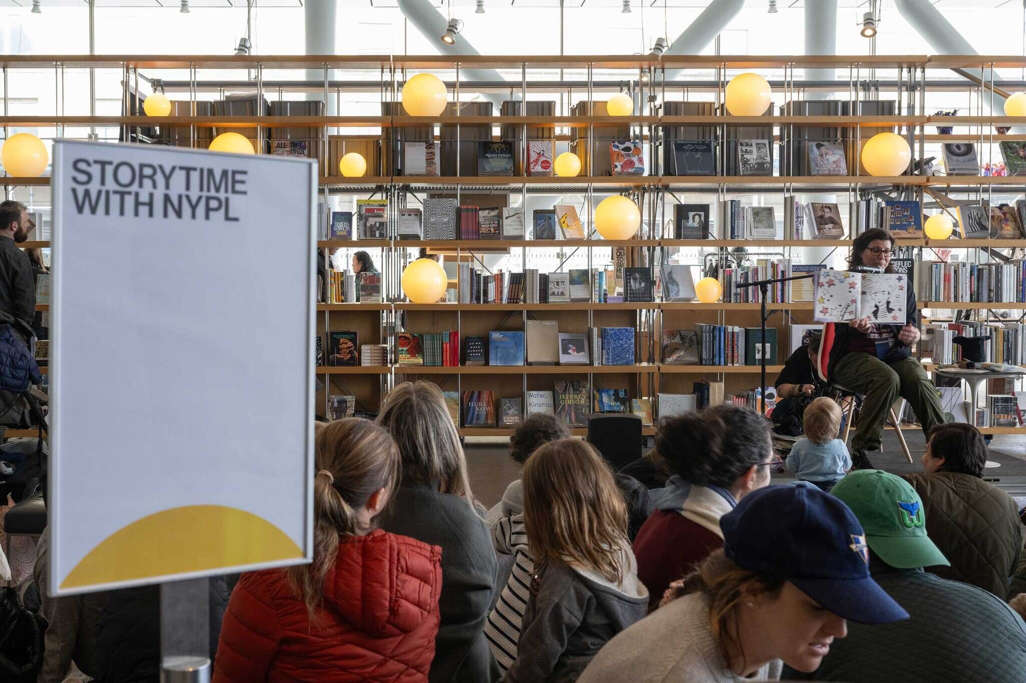 A group of children and adults sit in the lobby of the Whitney Museum, attentively listening to a Librarian reading a book. In the foreground, a sign reads 'Storytime with NYPL.' The background features bookshelves filled with books and warm overhead lighting.