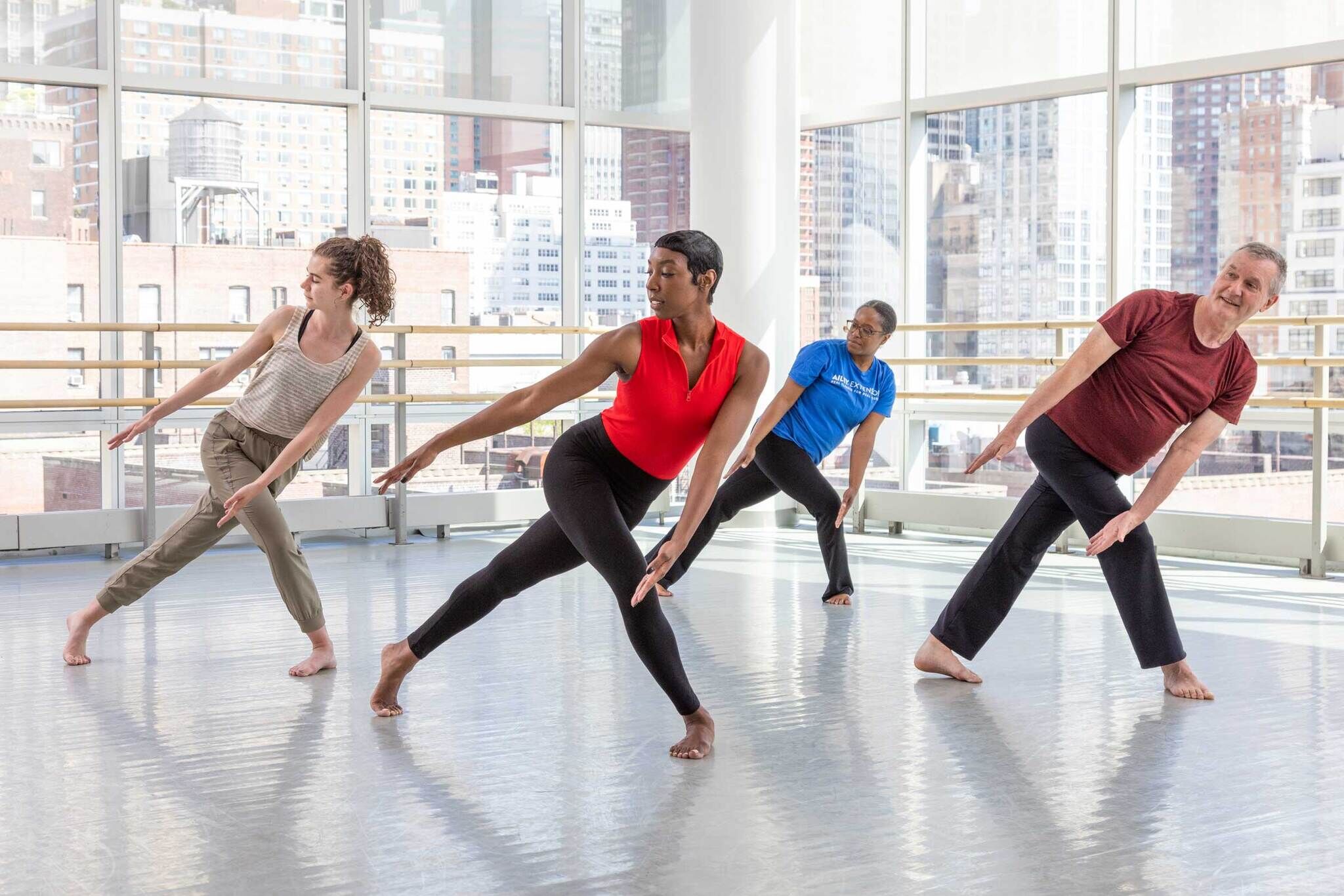 Four people stand in the corner of a light filled dance studio. They all stand with their back legs fully extended and their front legs bent across their bodies to form a triangle shape. Their arms are fully outstretched and reaching downwards and their faces are turned towards their back legs. The room has floor to ceiling windows that look out on nearby buildings in midtown Manhattan.