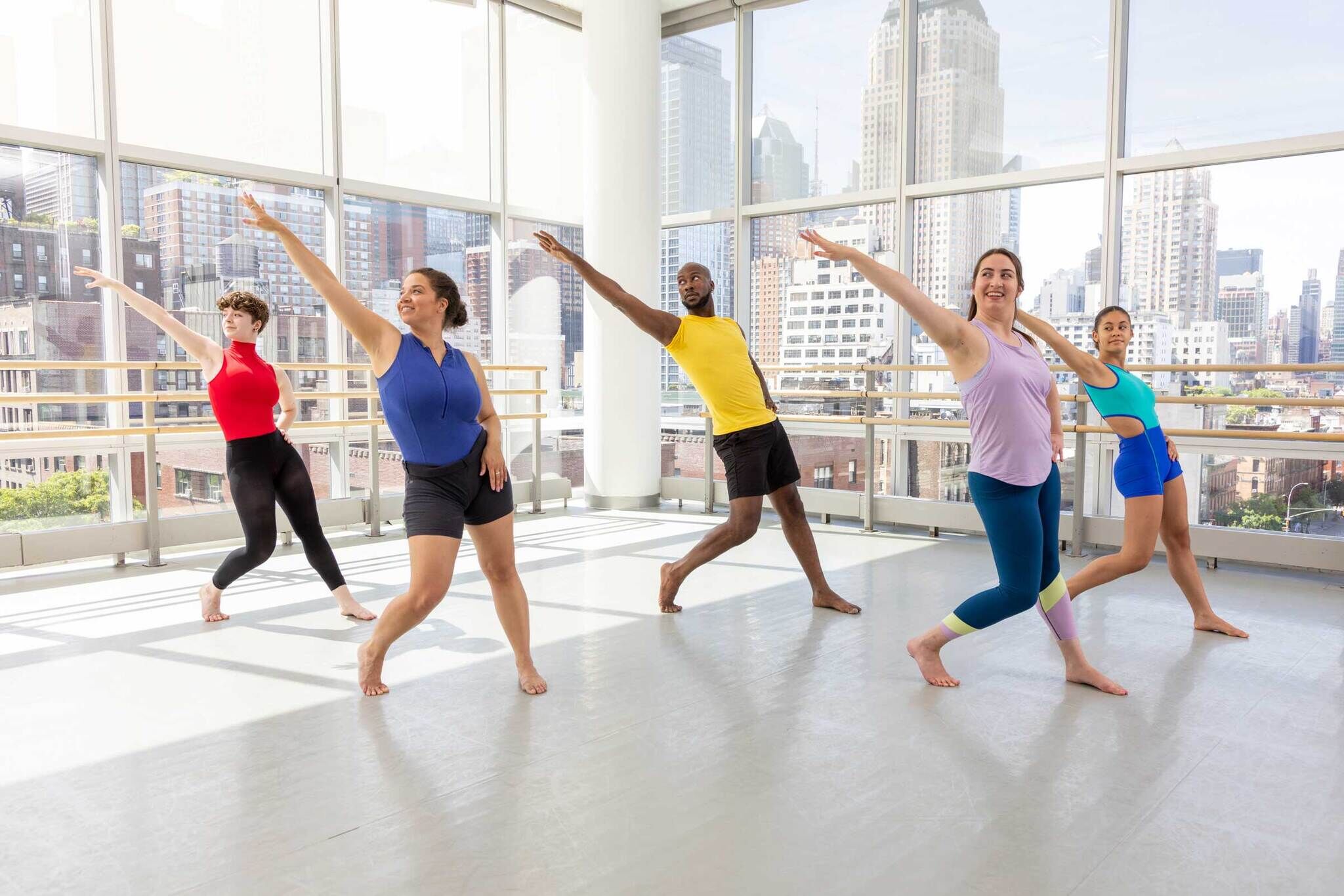 A group of five individuals in colorful exercise attire practice dance moves in a spacious studio with large windows showcasing a city skyline.