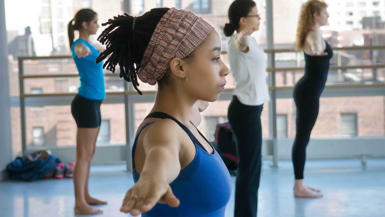 A group of four people stand in a dance studio, stretching their arms out to the sides. They are dressed in athletic wear and face forward in a line. The studio has large windows revealing an urban cityscape outside. The lead person wears a blue top and a headscarf.