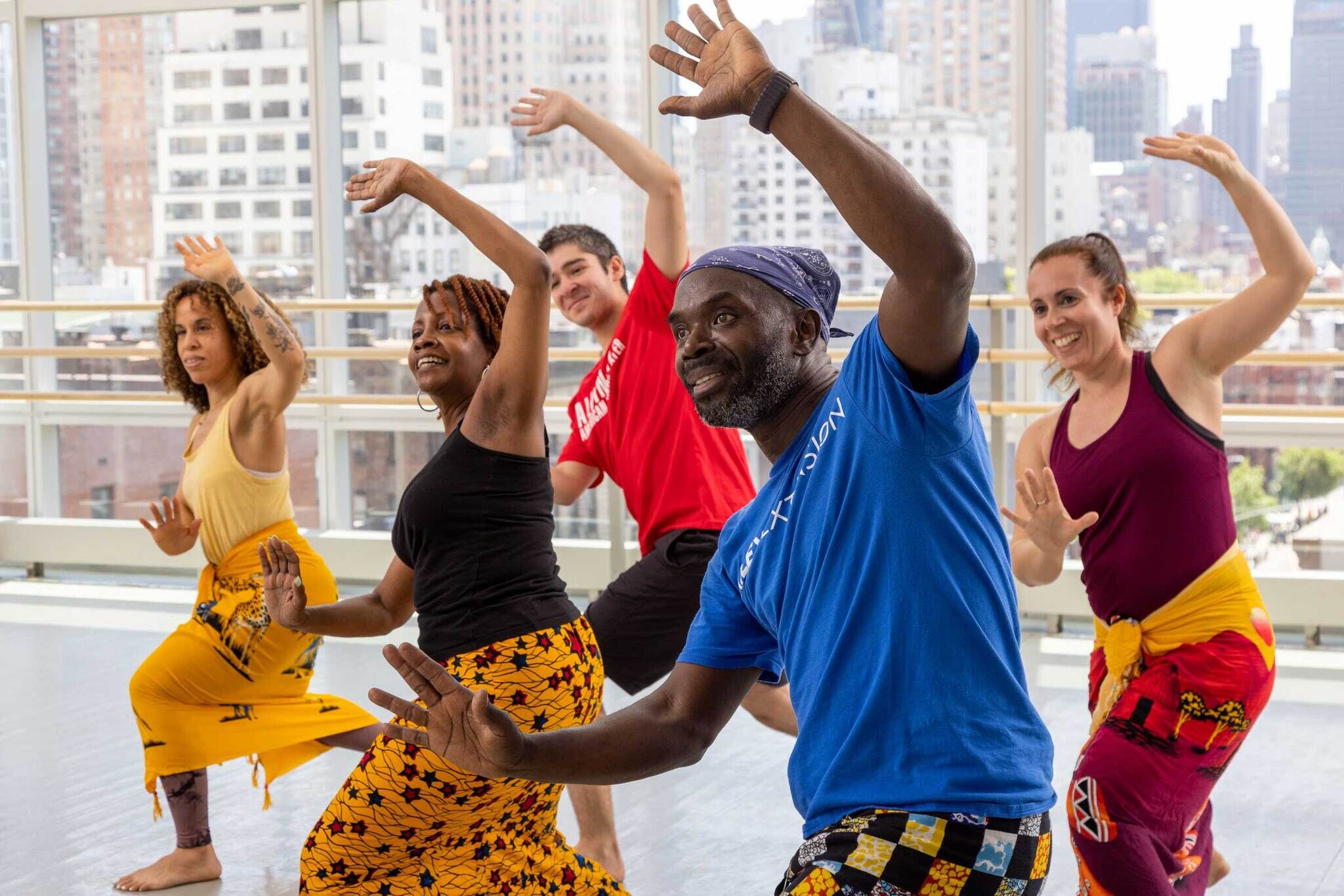 A diverse group of five people in colorful attire are dancing joyfully in a bright, spacious studio with large windows showing a cityscape. Everyone is smiling and has one arm raised, with the other arm bent at the elbow. The background includes tall buildings and a clear sky.