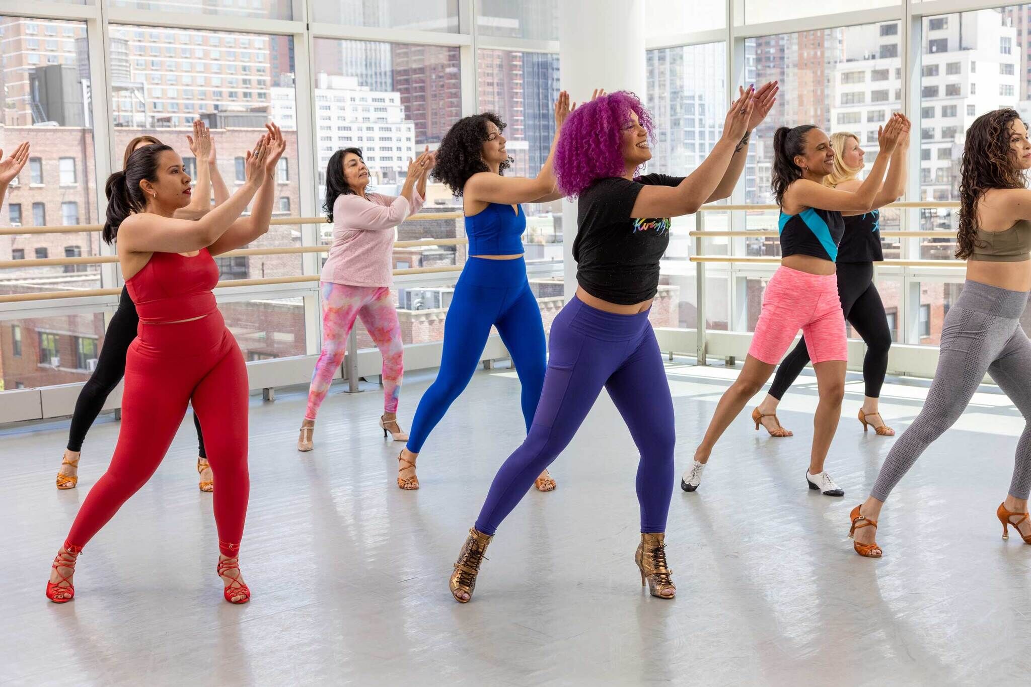 A group of women in colorful workout outfits are dancing together in a studio with large windows showing a cityscape. They appear synchronized and joyful, wearing high heels. The sunlight streams in, creating a bright and energetic atmosphere.