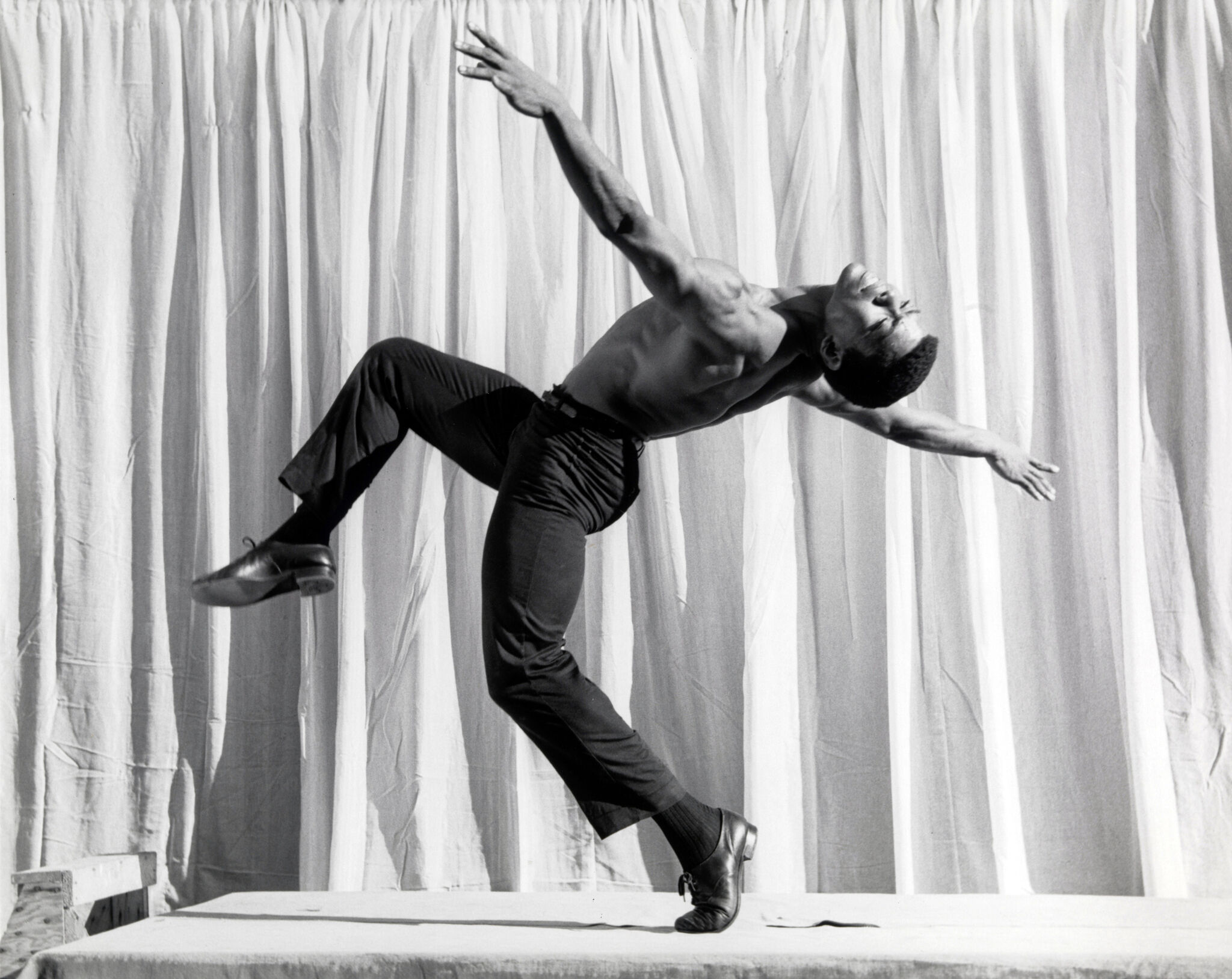 A shirtless man in black pants and shoes performs a dynamic dance pose against a backdrop of white curtains.