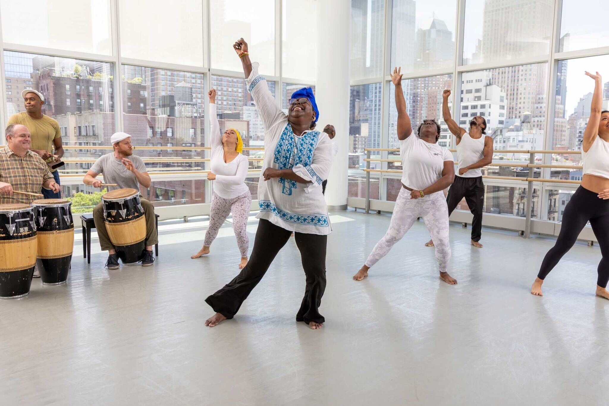 A group of people perform a dance routine in a bright studio with large windows showing a cityscape. One woman in the center wearing a blue headscarf and blue-and-white top leads the dance, while others around her follow. Three drummers on the left play traditional drums, providing the rhythm.