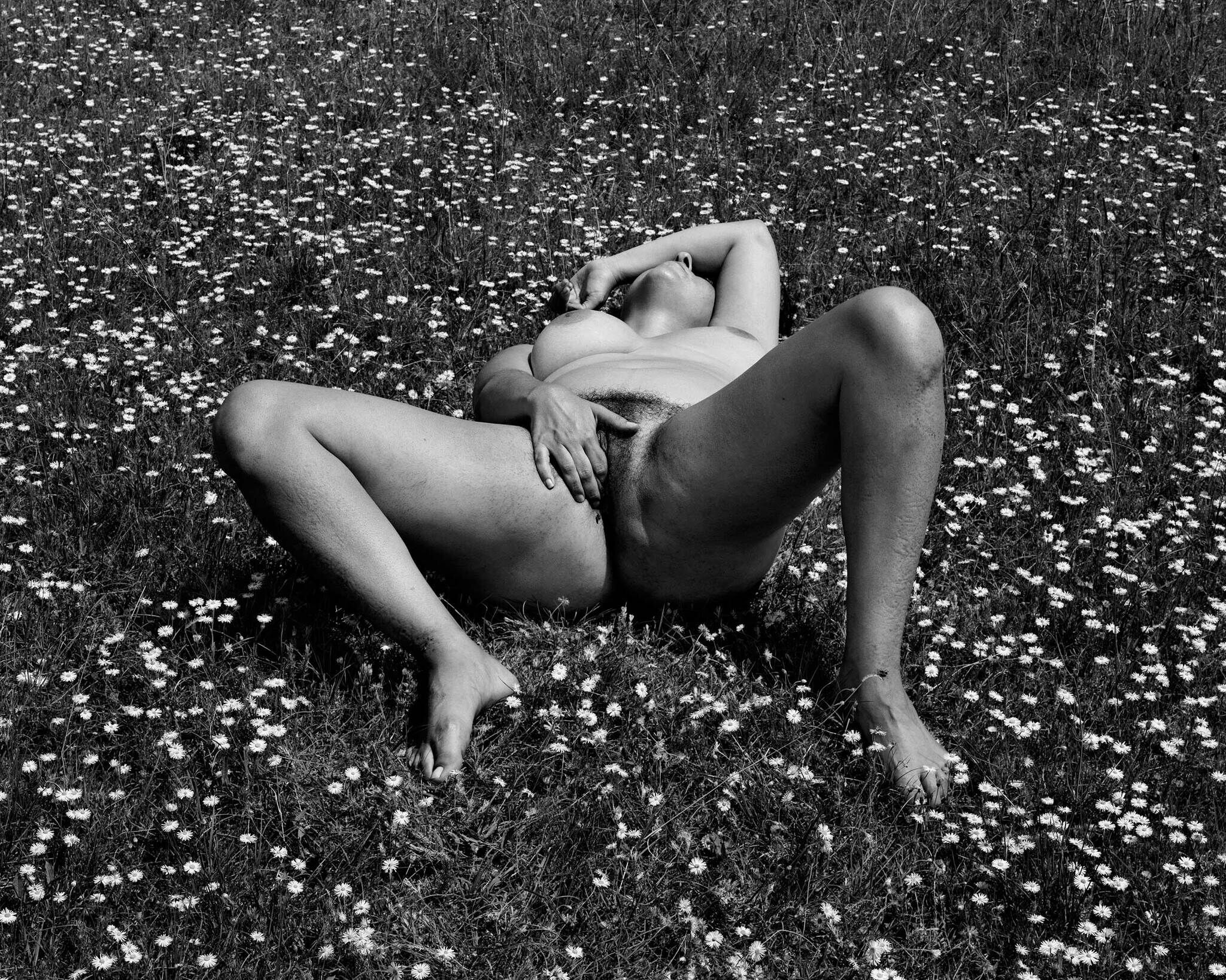 A naked person lies on their back in a field of flowers, legs spread and one hand covering their face, in a black and white photograph.