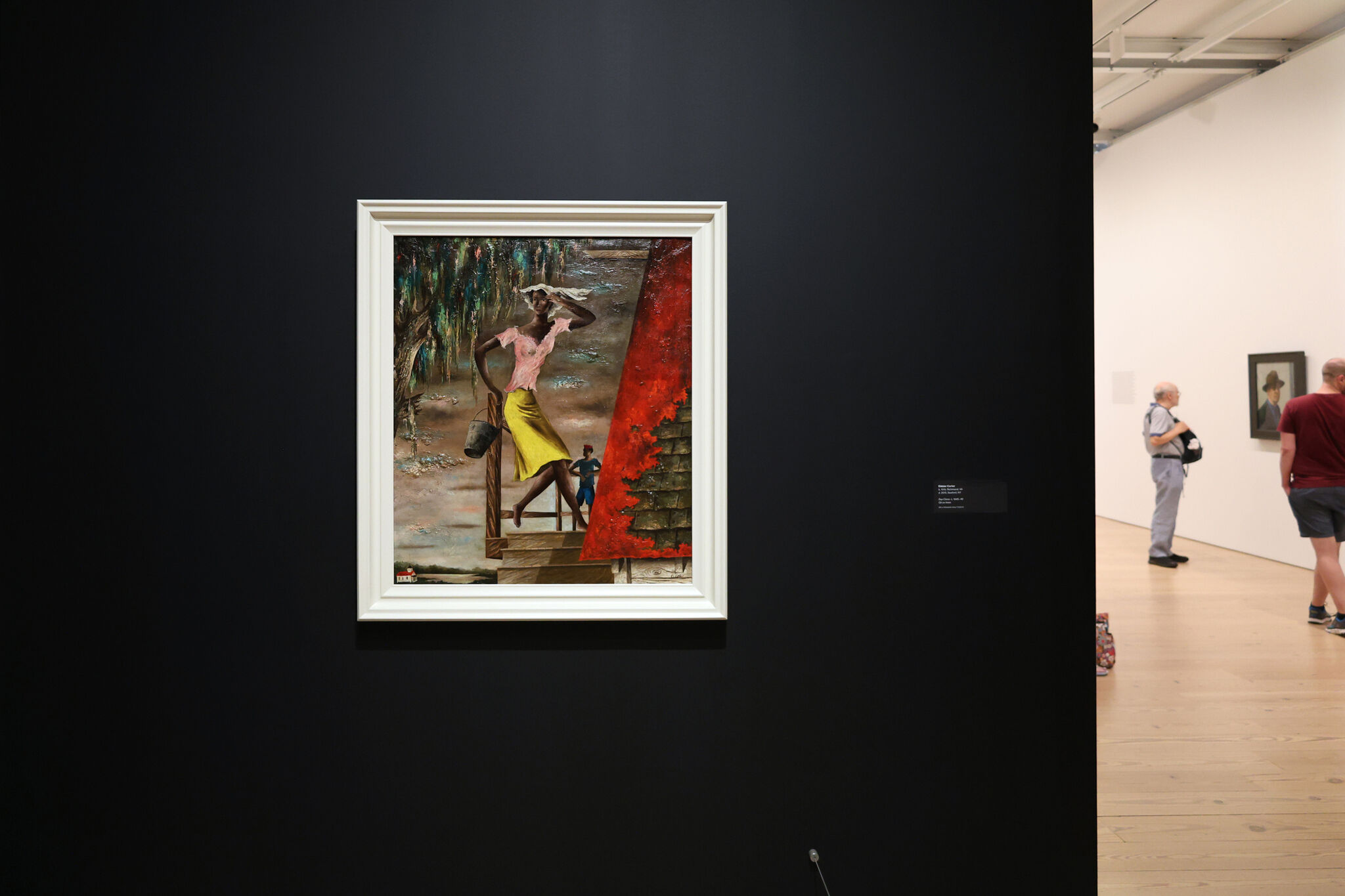 Painting of a woman in a yellow skirt and pink top, holding a bucket, displayed on a dark wall in an art gallery. Visitors observe other artworks.