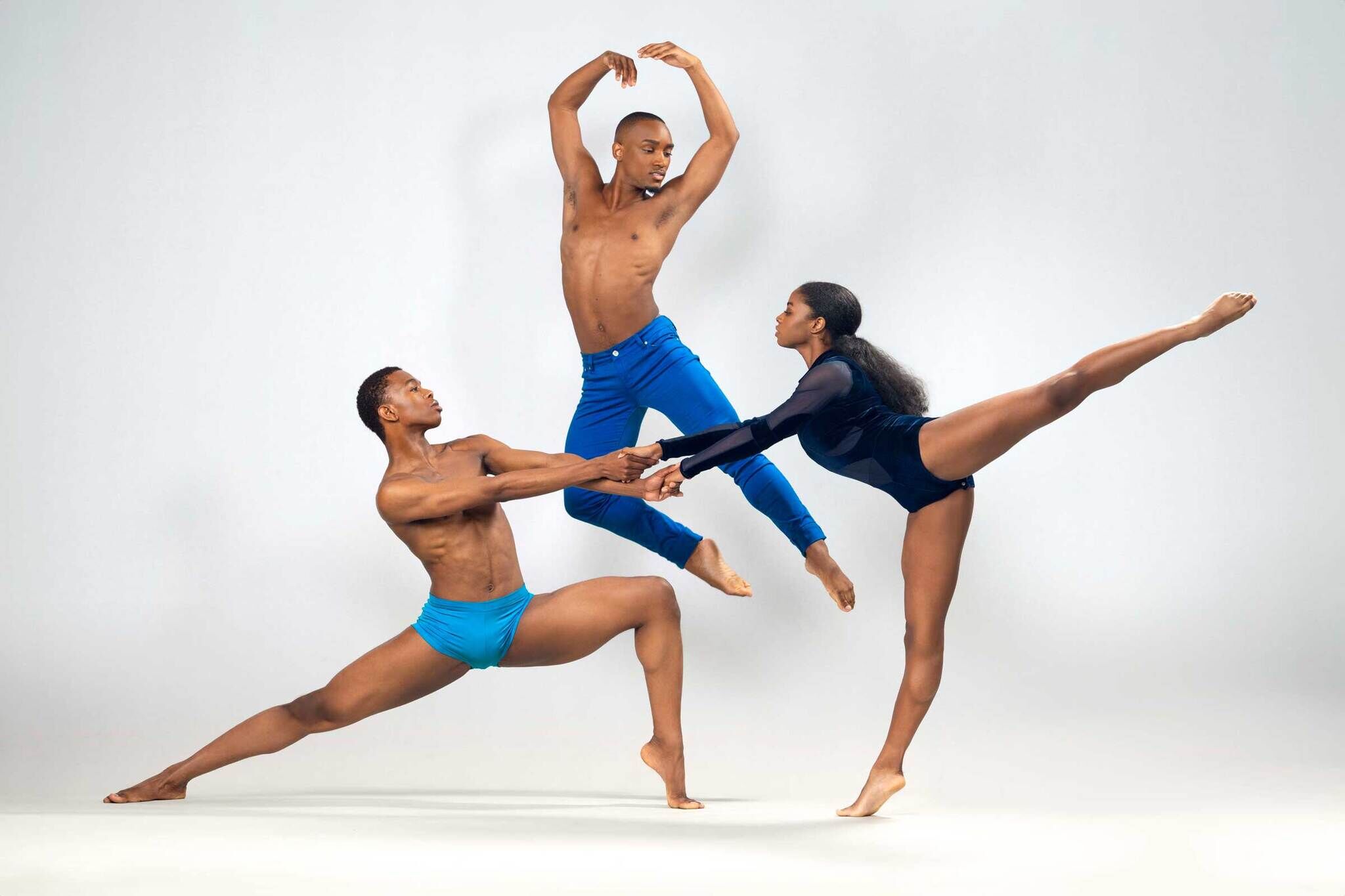 Three dancers in blue perform a dynamic pose: one leaps, another lunges, and the third extends a leg high, all holding hands.