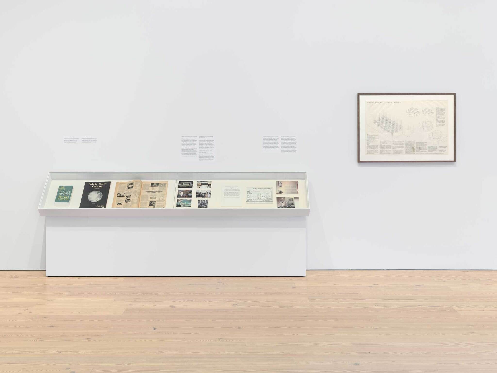 Museum display with books and documents in a glass case, and a framed diagram on the wall. Text descriptions are mounted on the white wall.